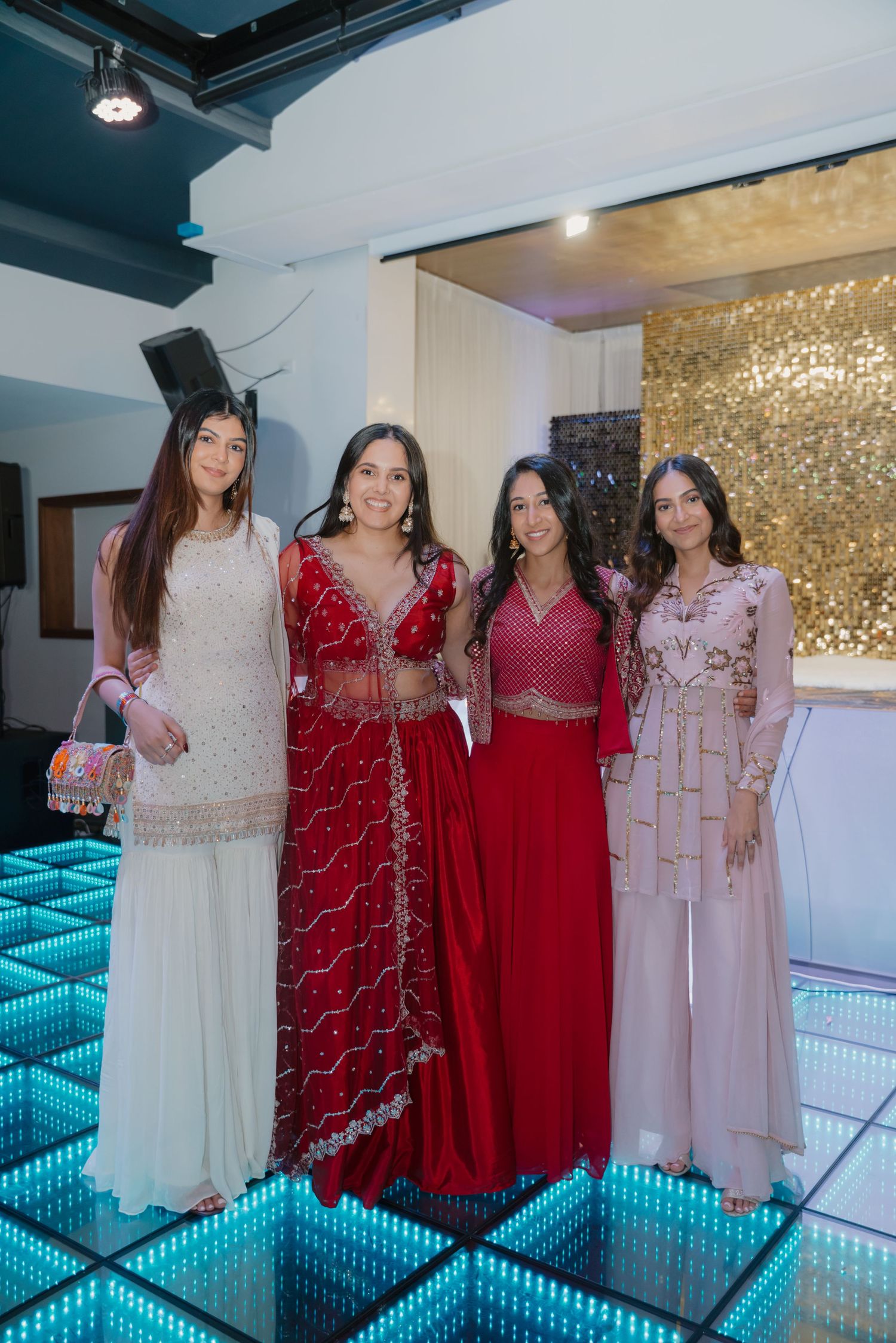 Four women in elegant Indian ethnic wear pose on LED dance floor at wedding reception.