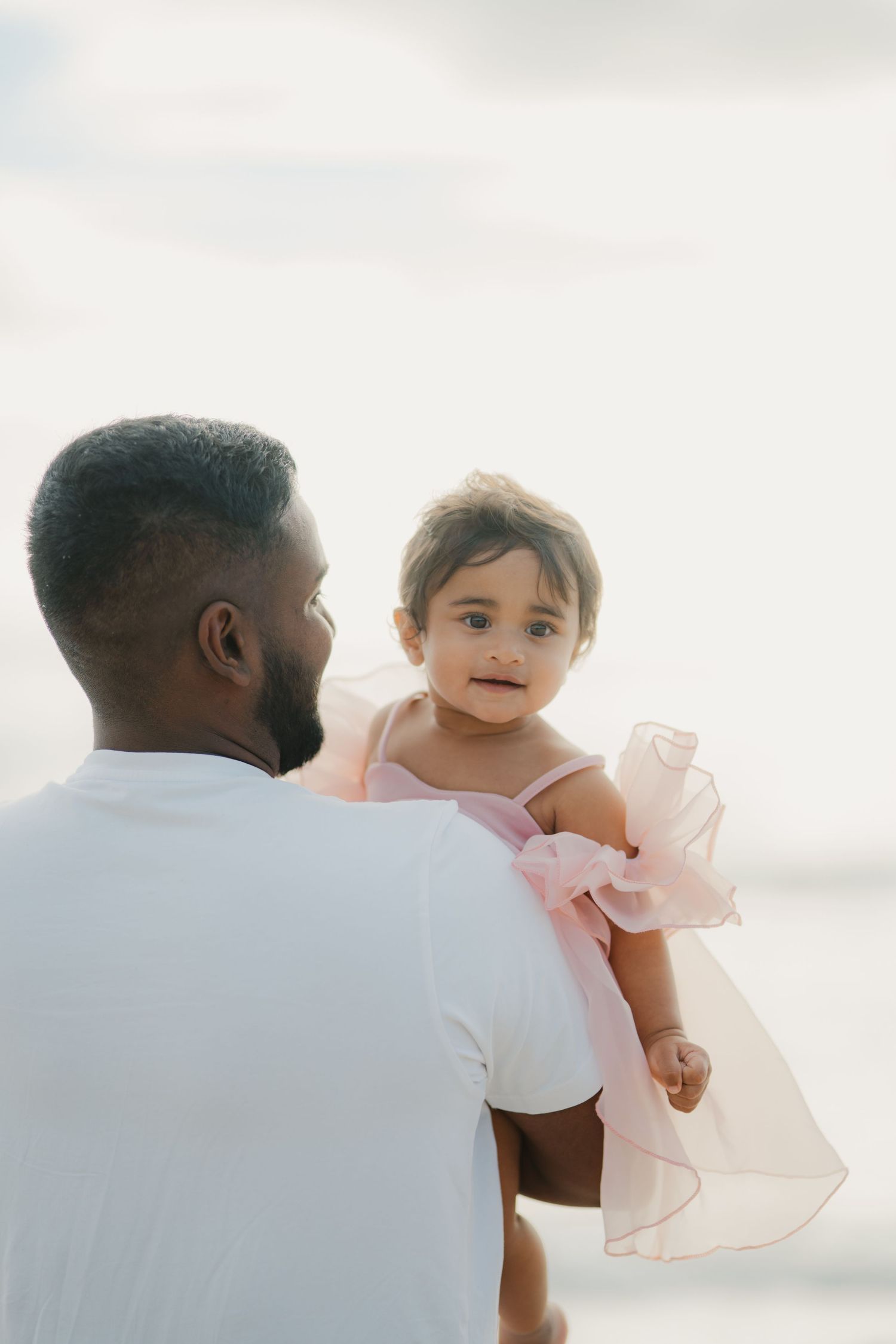 Father in white shirt holding his baby girl in a pink dress on a bright beach.