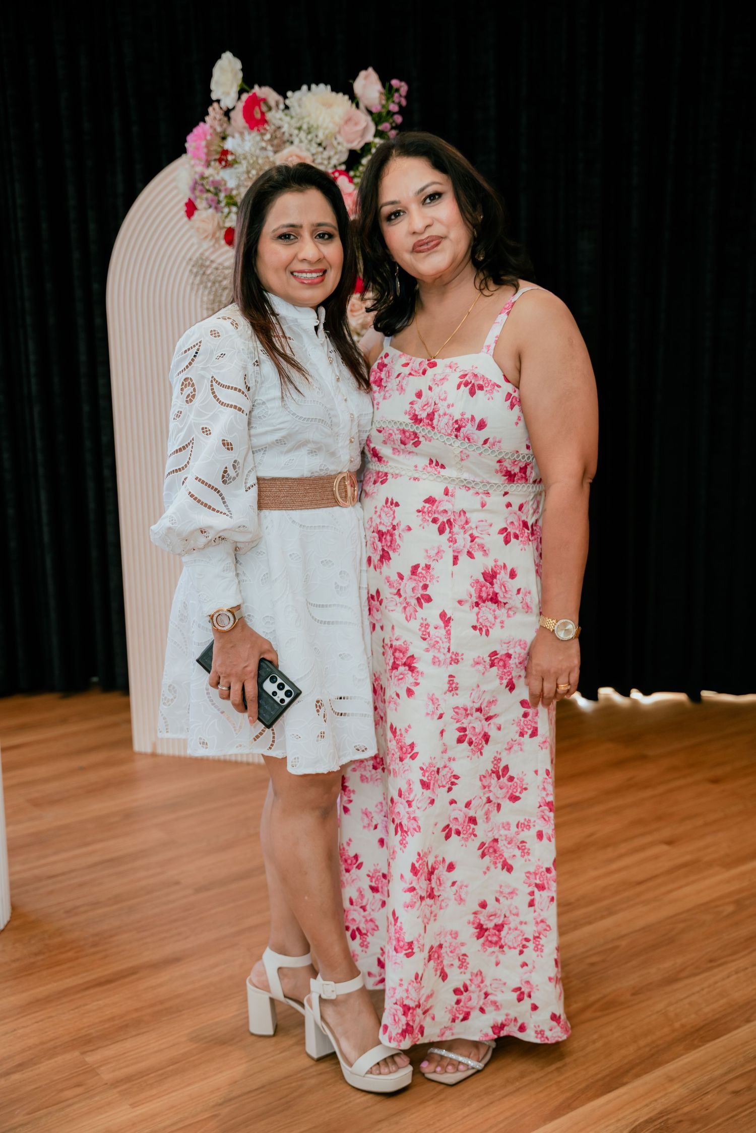 Two women in floral and white dresses smile together at an elegant birthday party event.