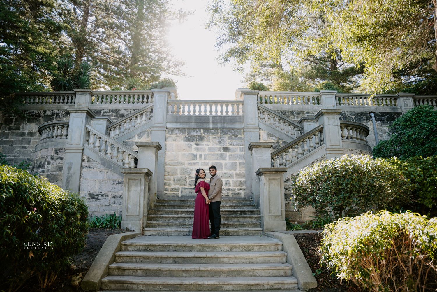Couple embracing on ornate stone staircase with balustrades and tall trees in soft sunlight.