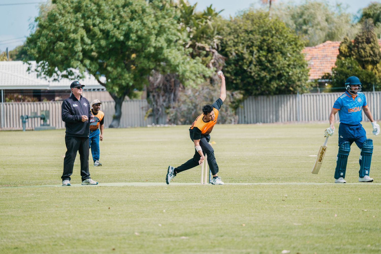 Cricket bowler in orange uniform releases ball during match on grass pitch.