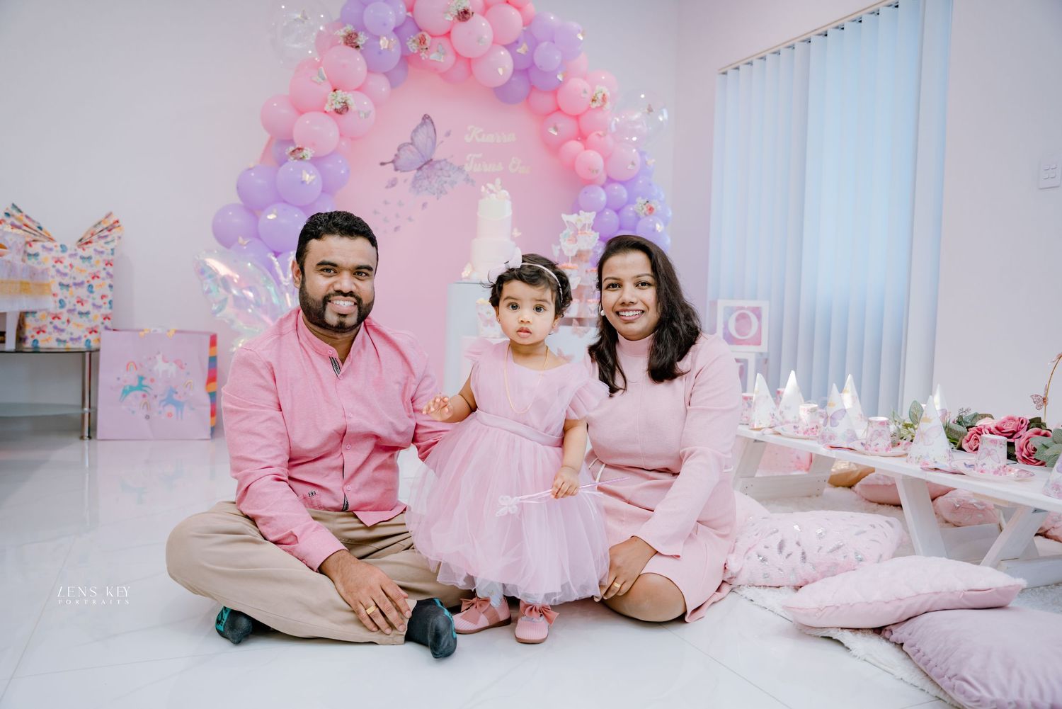 Family of three sitting on floor at pink butterfly-themed first birthday party with balloon decorations.