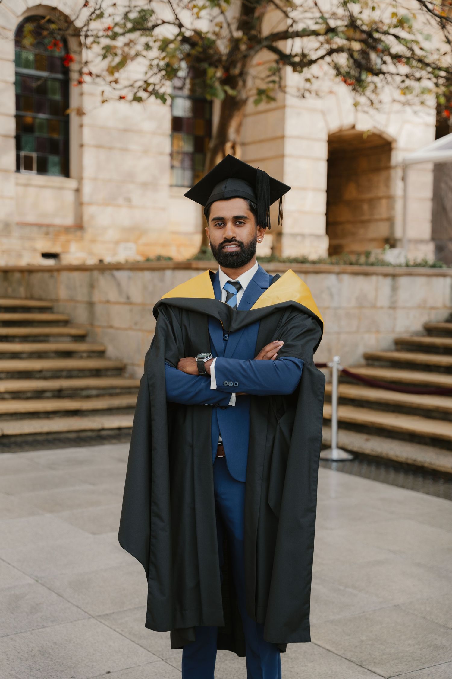 Male graduate in blue suit and black gown with gold hood stands arms crossed outdoors.