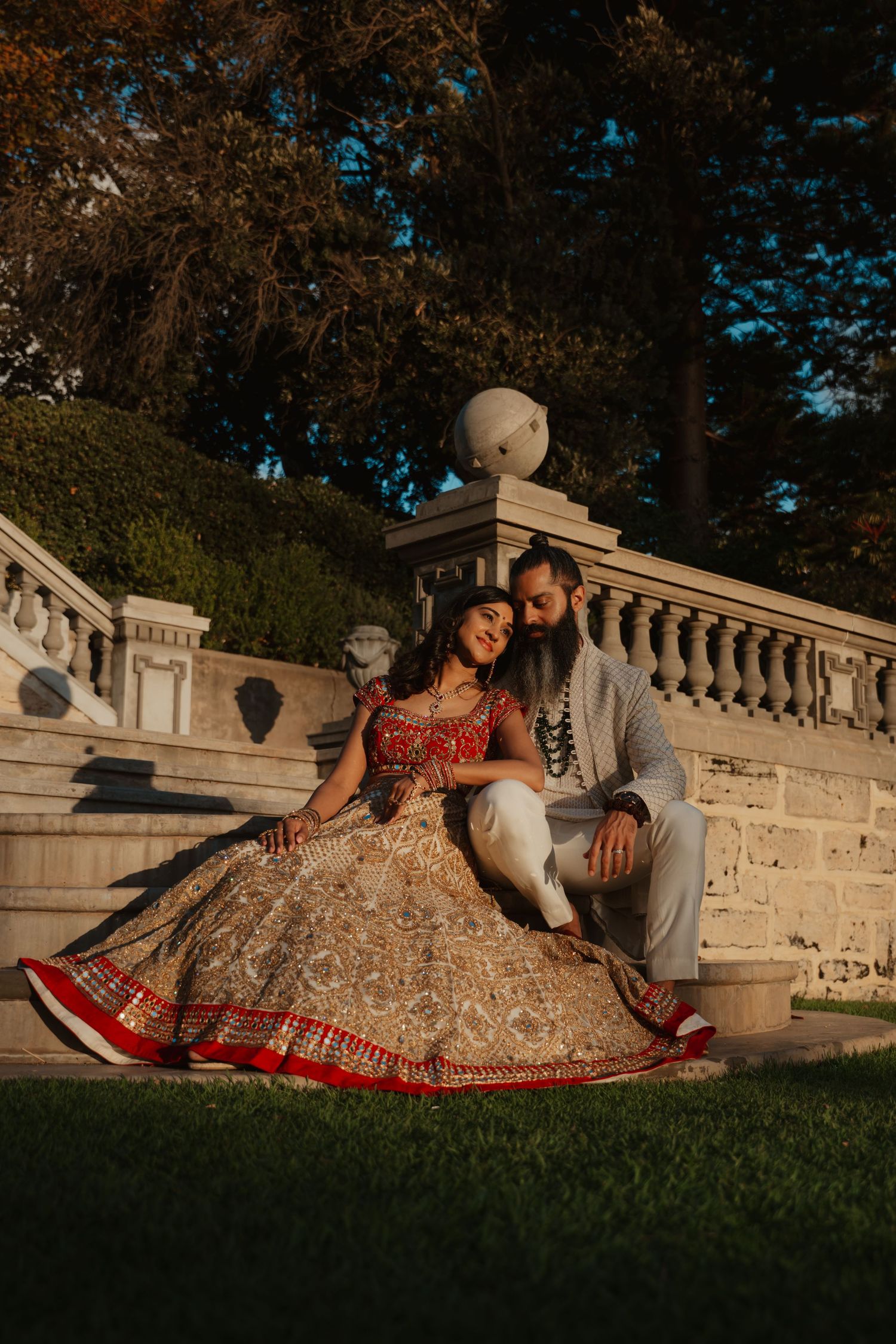 Indian wedding couple in traditional attire sit together on grass steps during warm sunset light.
