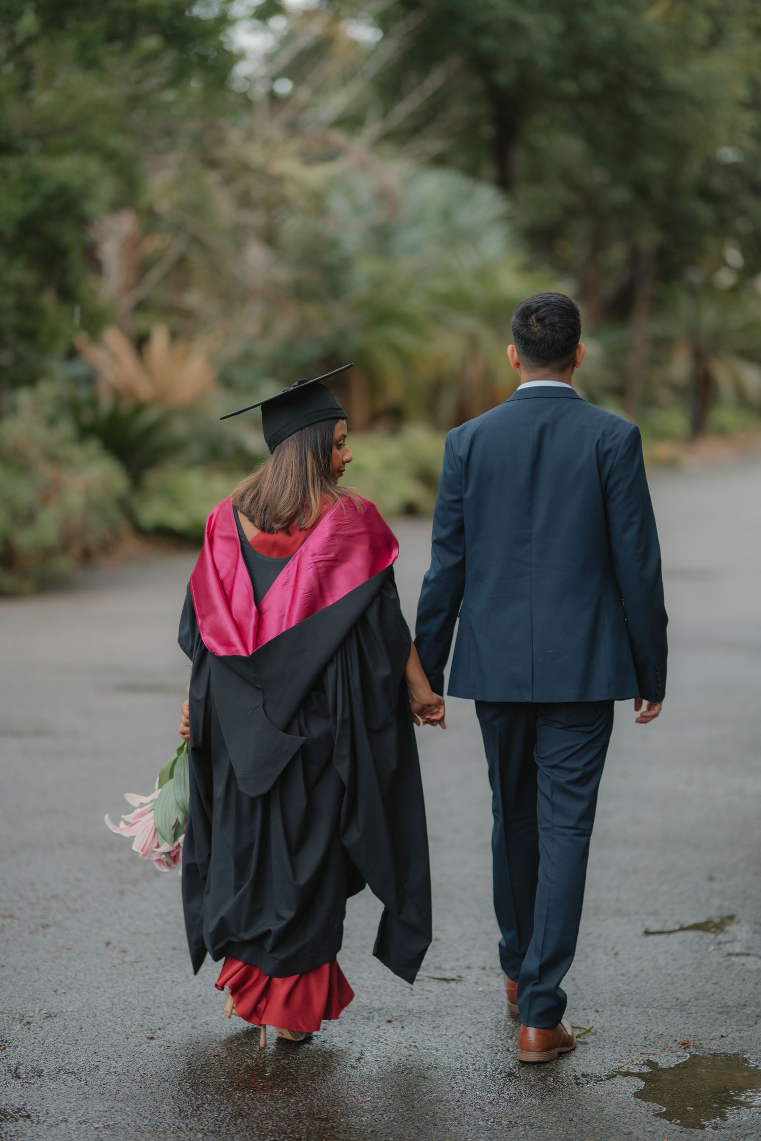 Rear view of graduate in black gown and red dress walking hand-in-hand with partner on wet path.
