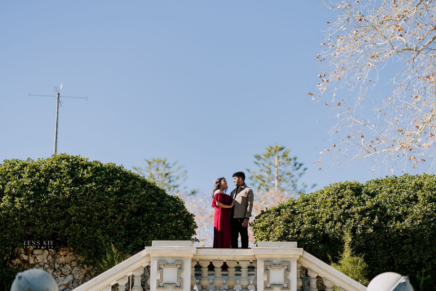 Couple standing on white stone steps surrounded by lush green hedges during an outdoor engagement photo session.