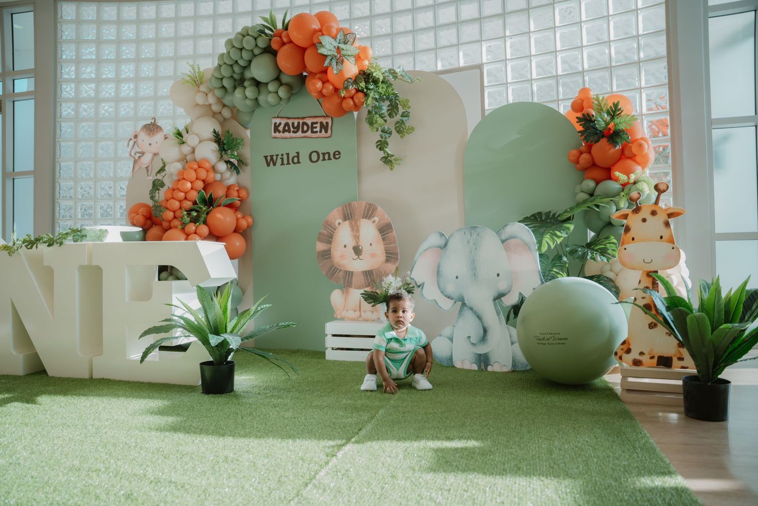 A baby sits on green turf at a Wild One safari birthday party with elephant and lion themed decorations.