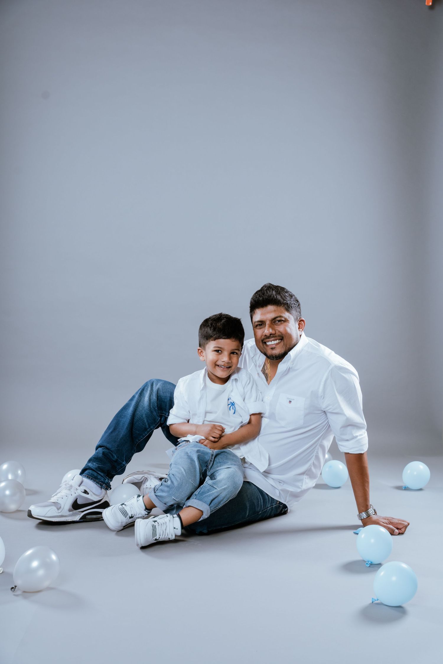 Father and son in matching white shirts sitting on floor with blue balloons in a studio photo shoot.