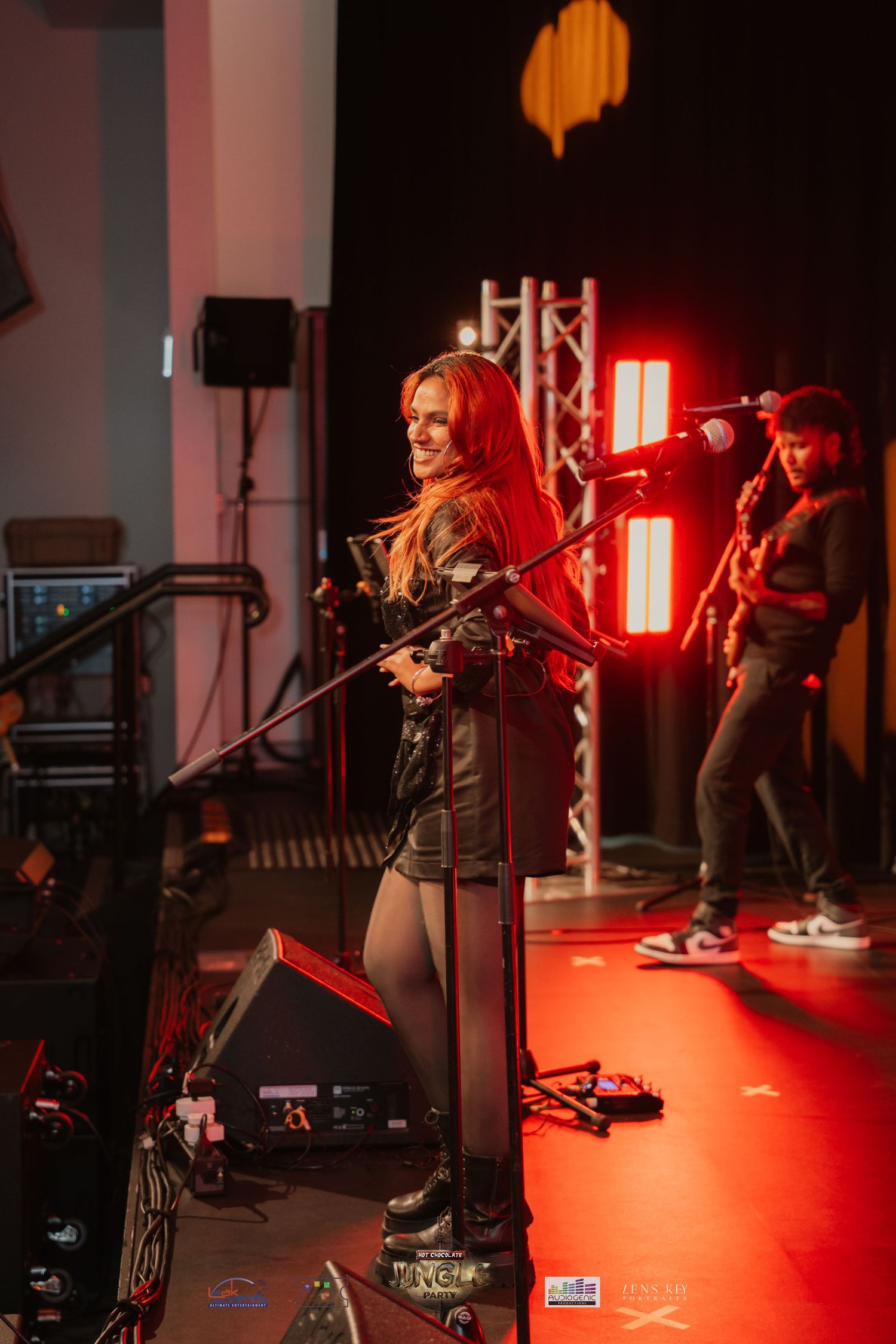 Female vocalist smiling on stage holding microphone stand under red lights with guitarist behind.