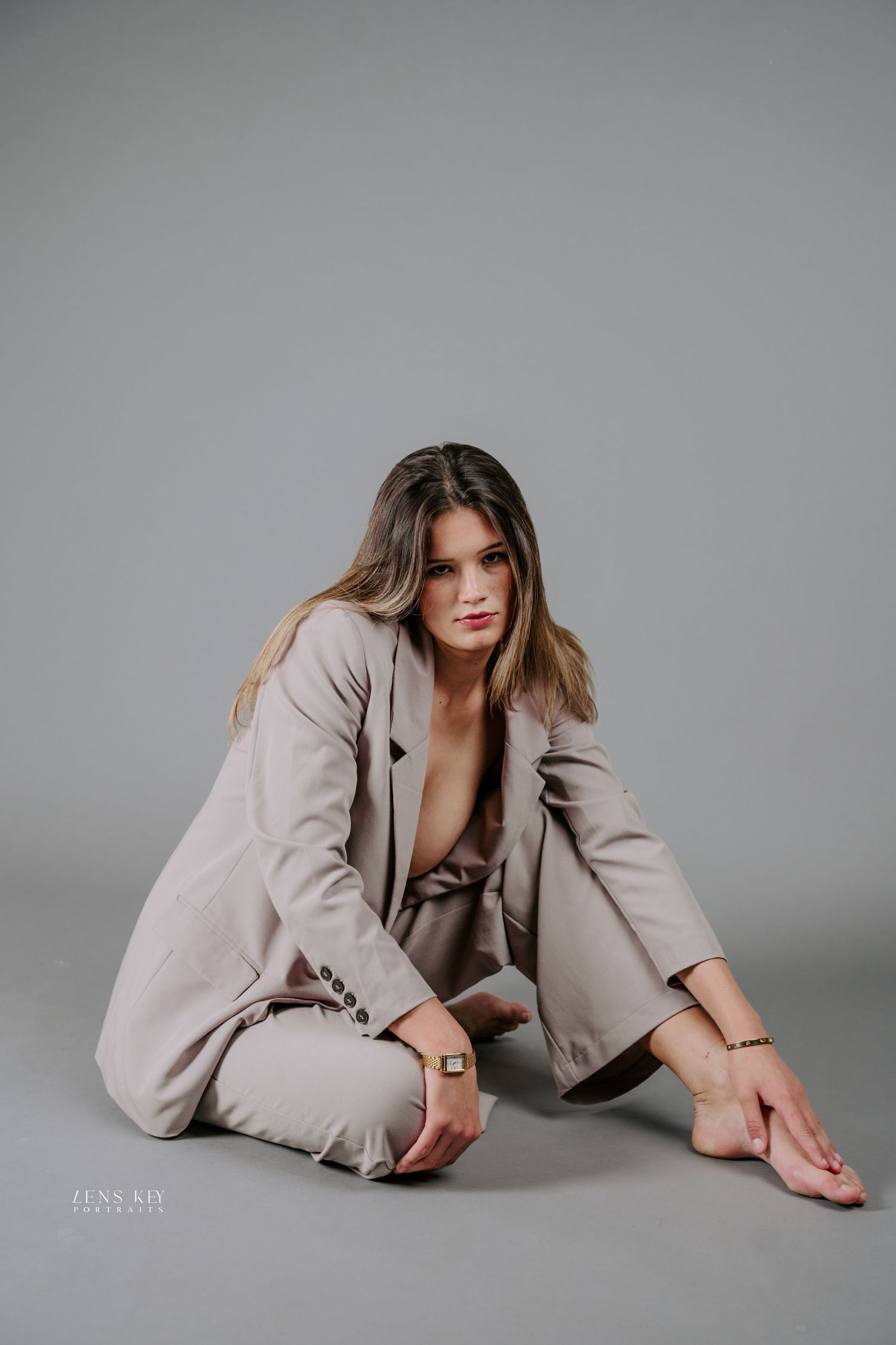 Woman in beige oversized blazer suit sitting on floor in elegant fashion portrait on gray background.