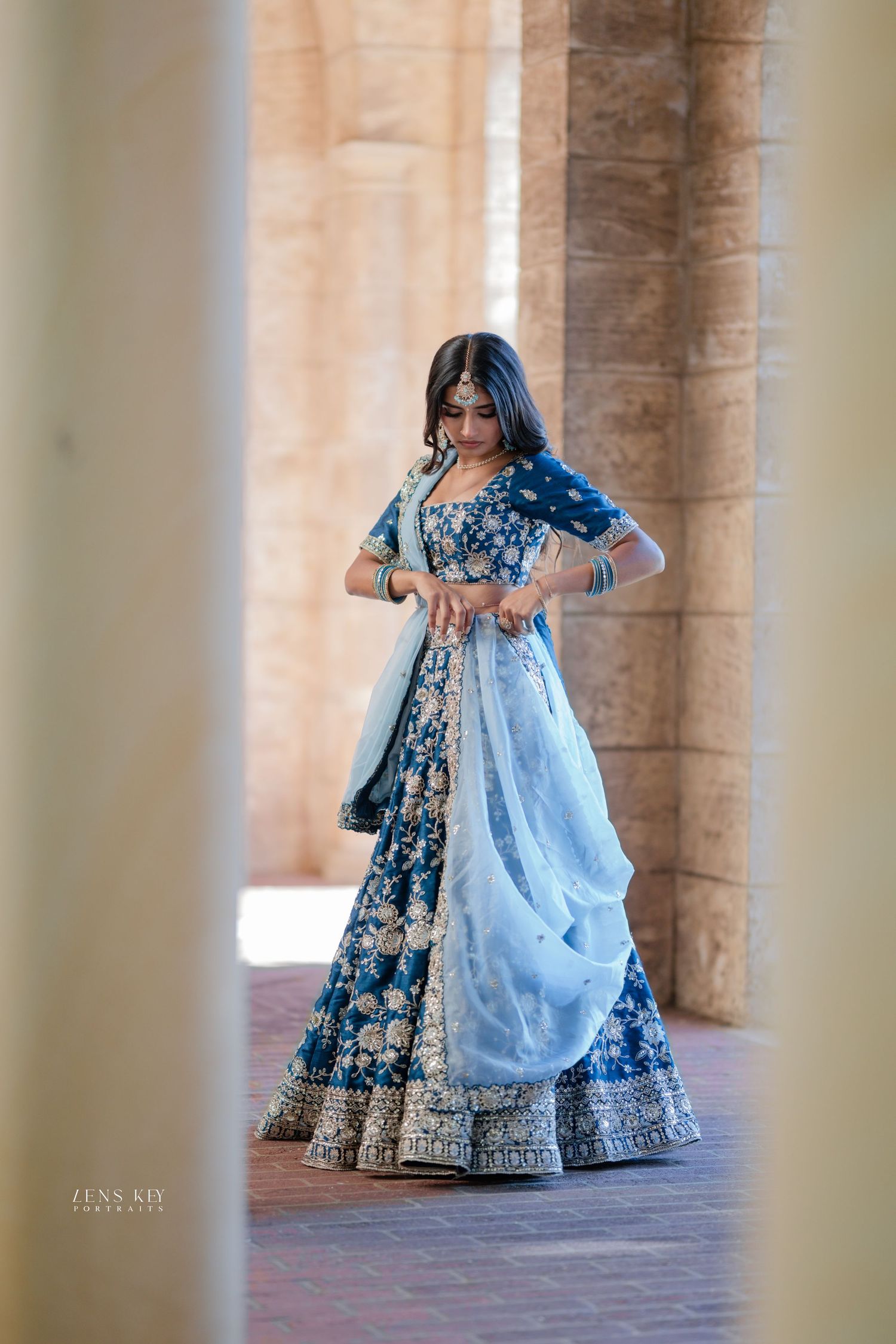 Woman in blue embroidered lehenga with light blue dupatta posing between stone columns.
