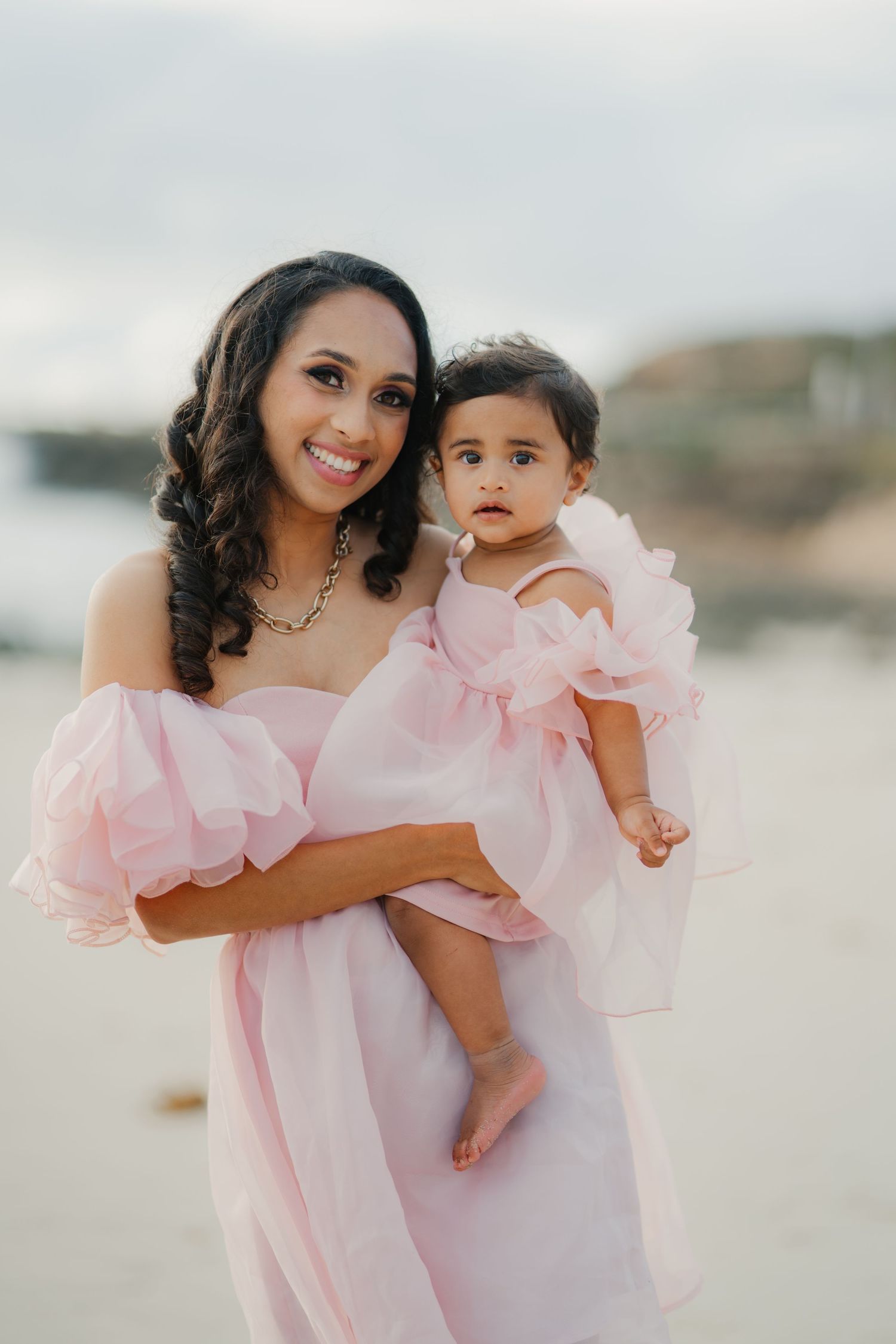 Mother and baby daughter in matching pink dresses posing together on a sandy beach.