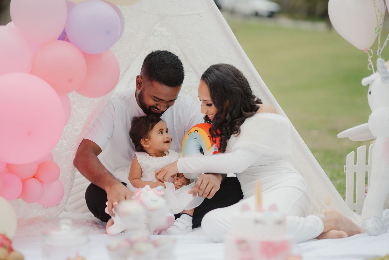 Parents lovingly attending to baby at whimsical outdoor birthday party with pastel balloons and teepee backdrop.