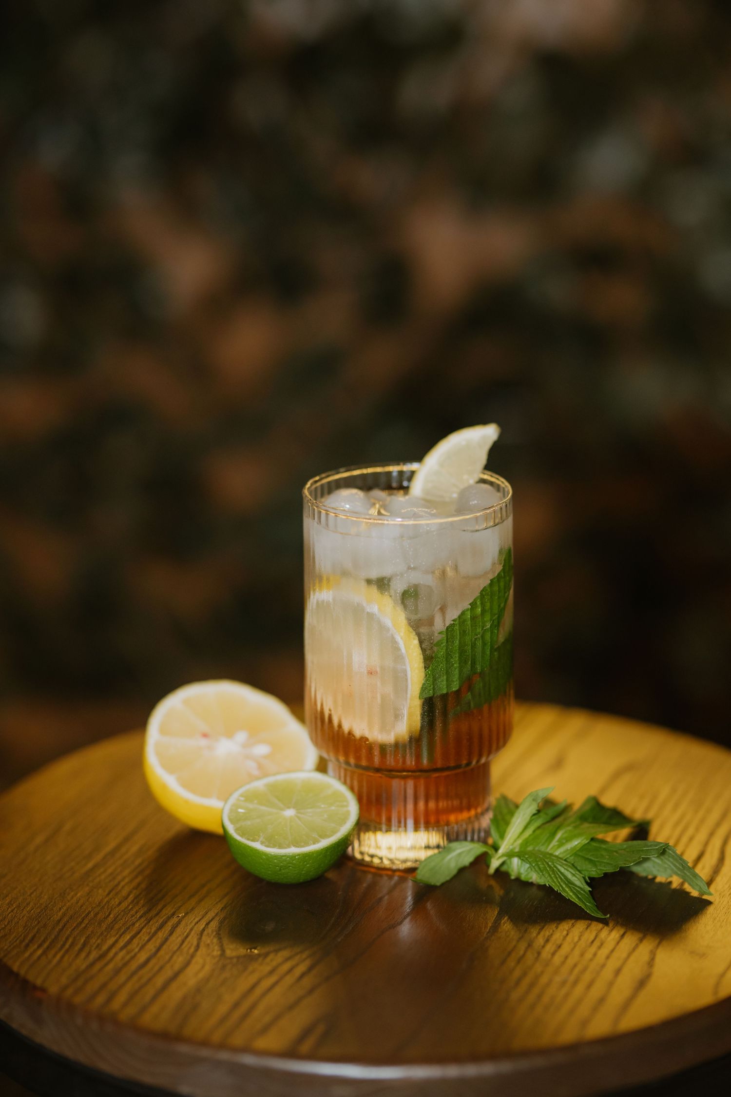 Iced tea cocktail with lemon lime slices and fresh mint in tall glass on wooden table.