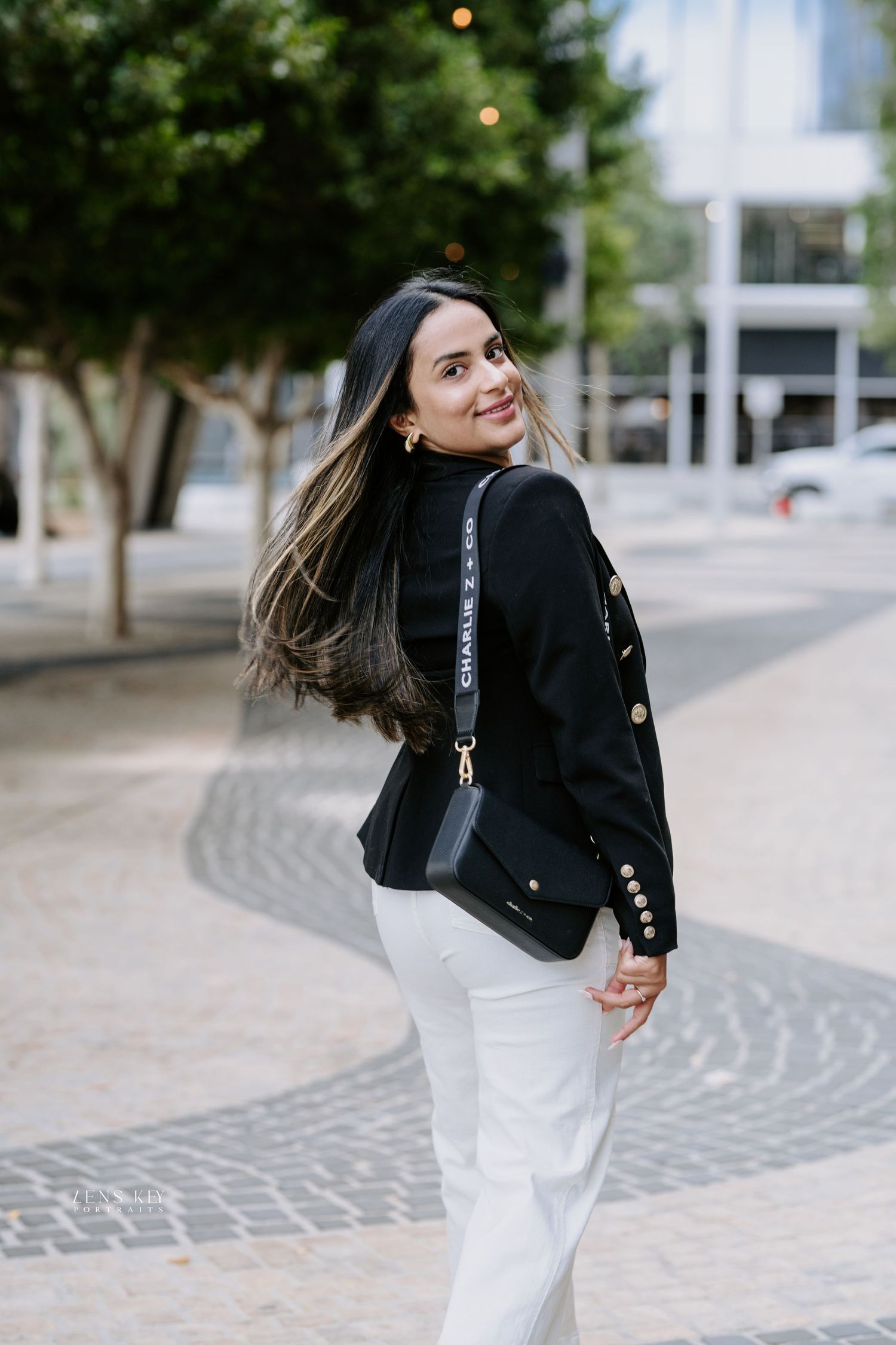 Woman in black blazer white pants smiling over shoulder on decorative cobblestone plaza.