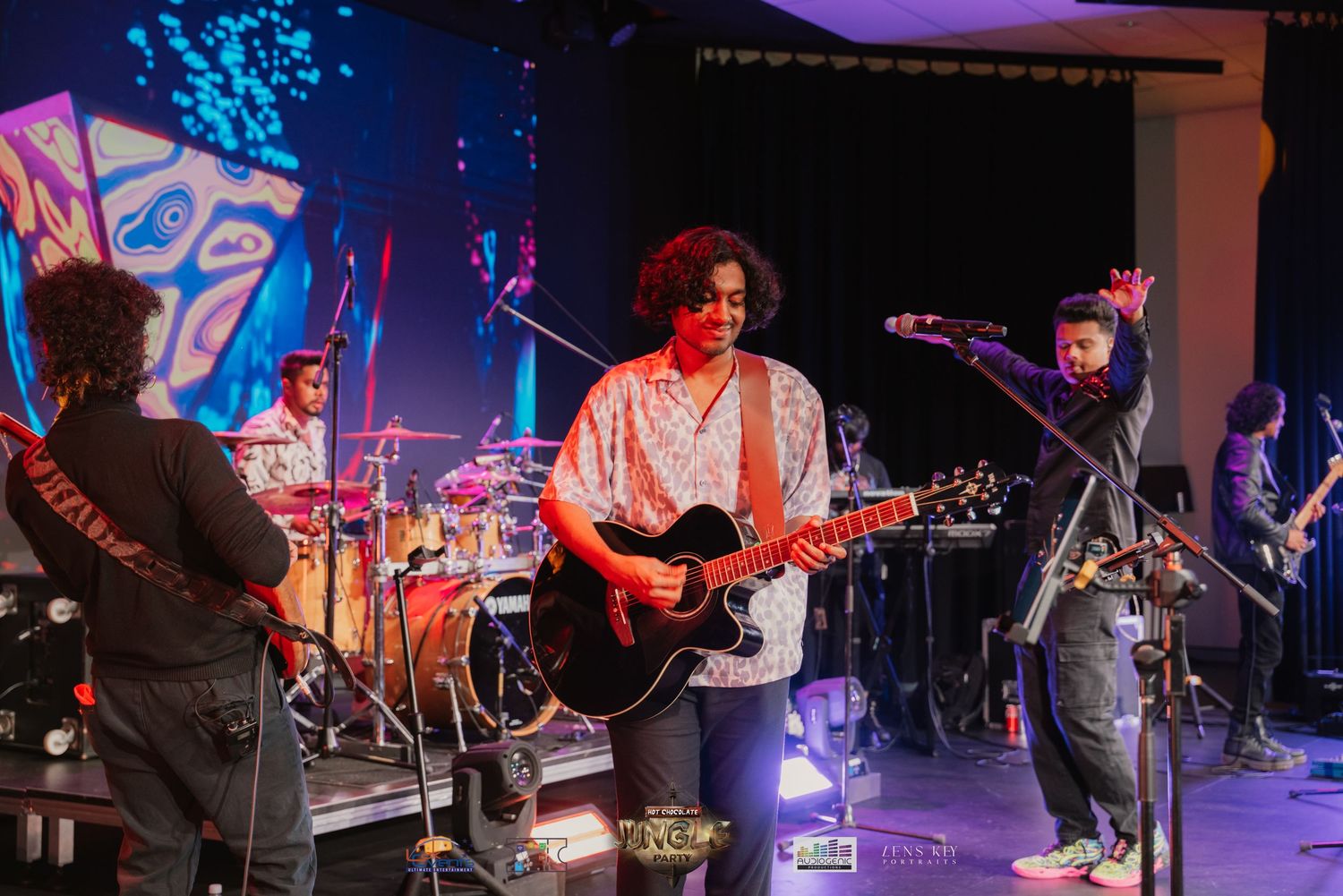 Band performing on stage with guitarist in floral shirt under colorful lights with artistic backdrop.