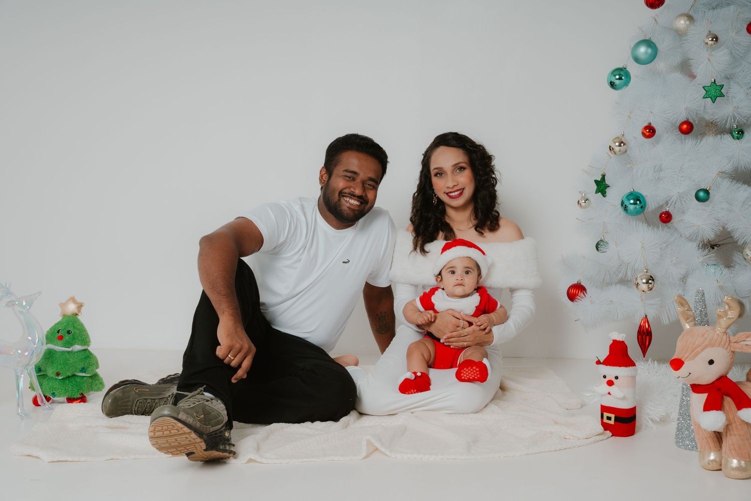 Smiling family with infant in Santa costume posing beside a white Christmas tree with colorful ornaments.
