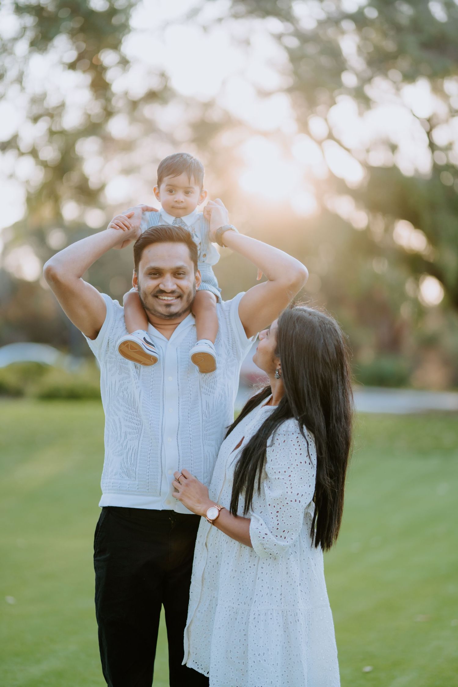 Parents in white outfits smile as dad carries toddler on shoulders at golden hour park.