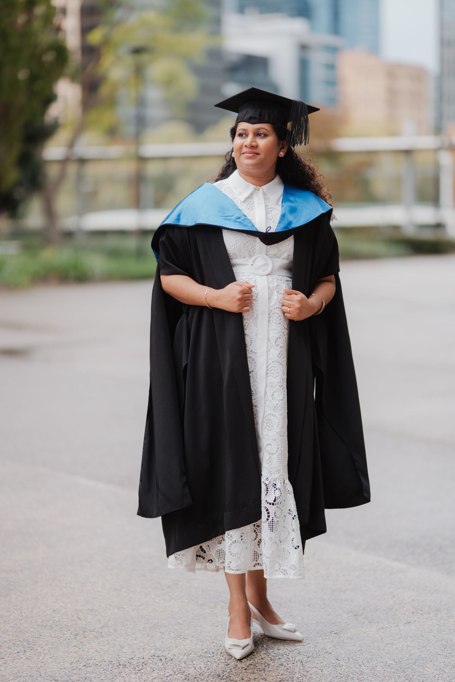 Graduate woman in black cap and gown with blue hood stands confidently on campus.