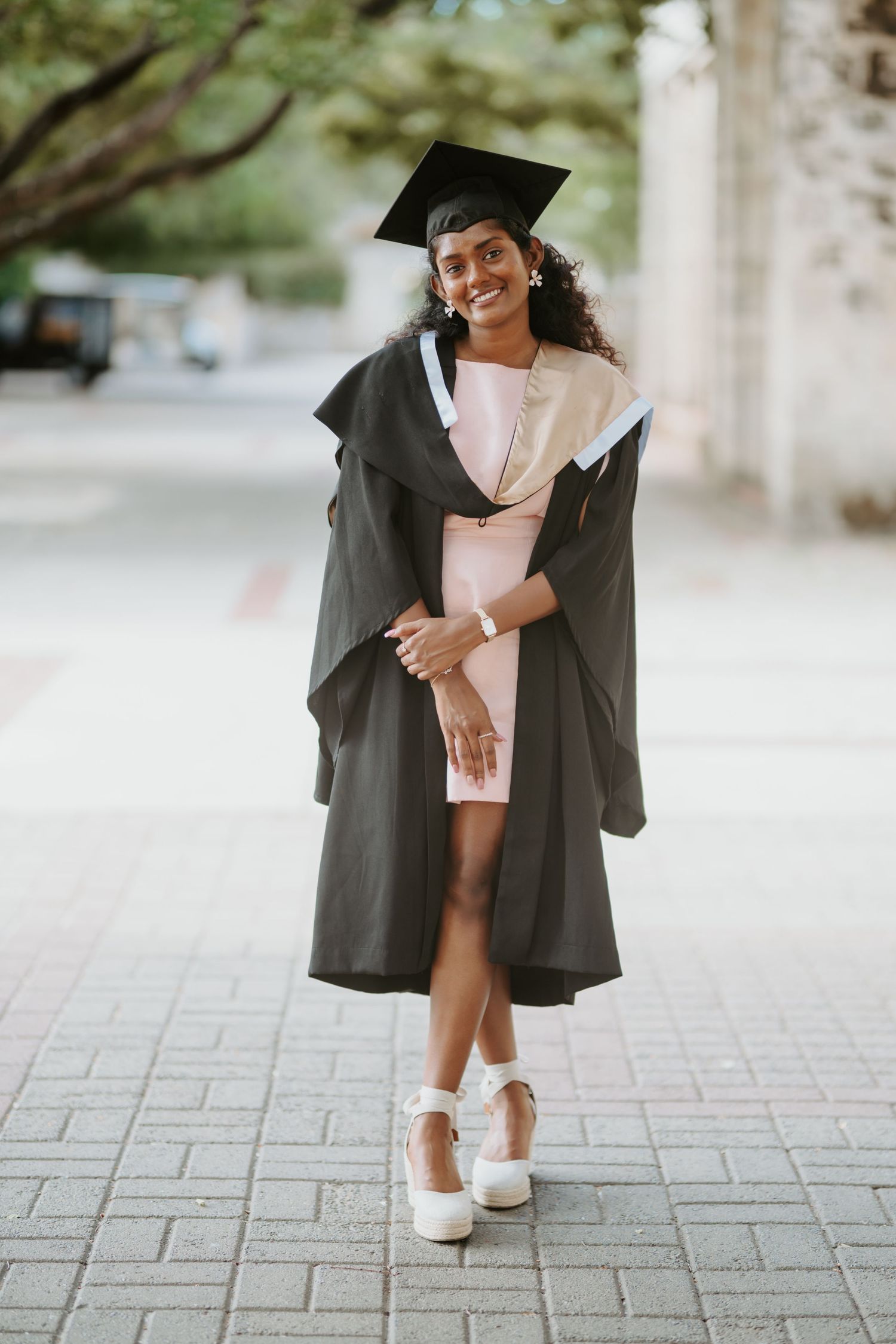 Smiling graduate in black gown and mortarboard cap poses outdoors on paved walkway.