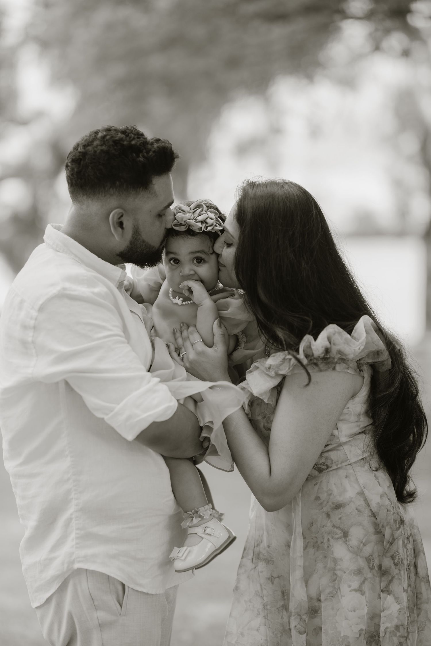 Black and white photo of parents lovingly kissing their baby girl outdoors in a park setting.