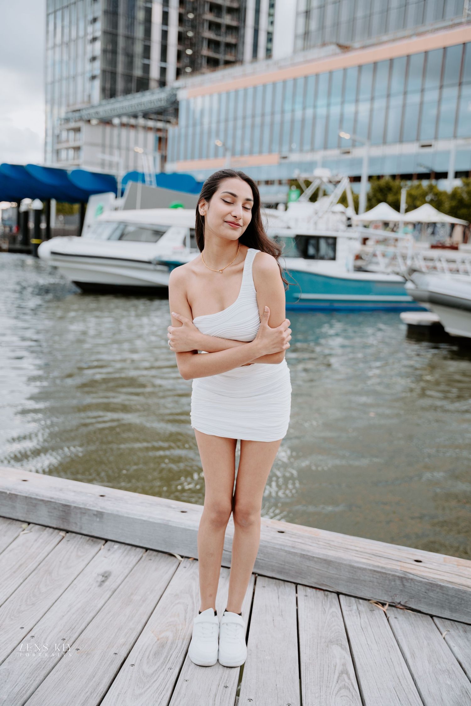 Young woman in white one-shoulder mini dress stands on wooden dock with marina and boats behind her.