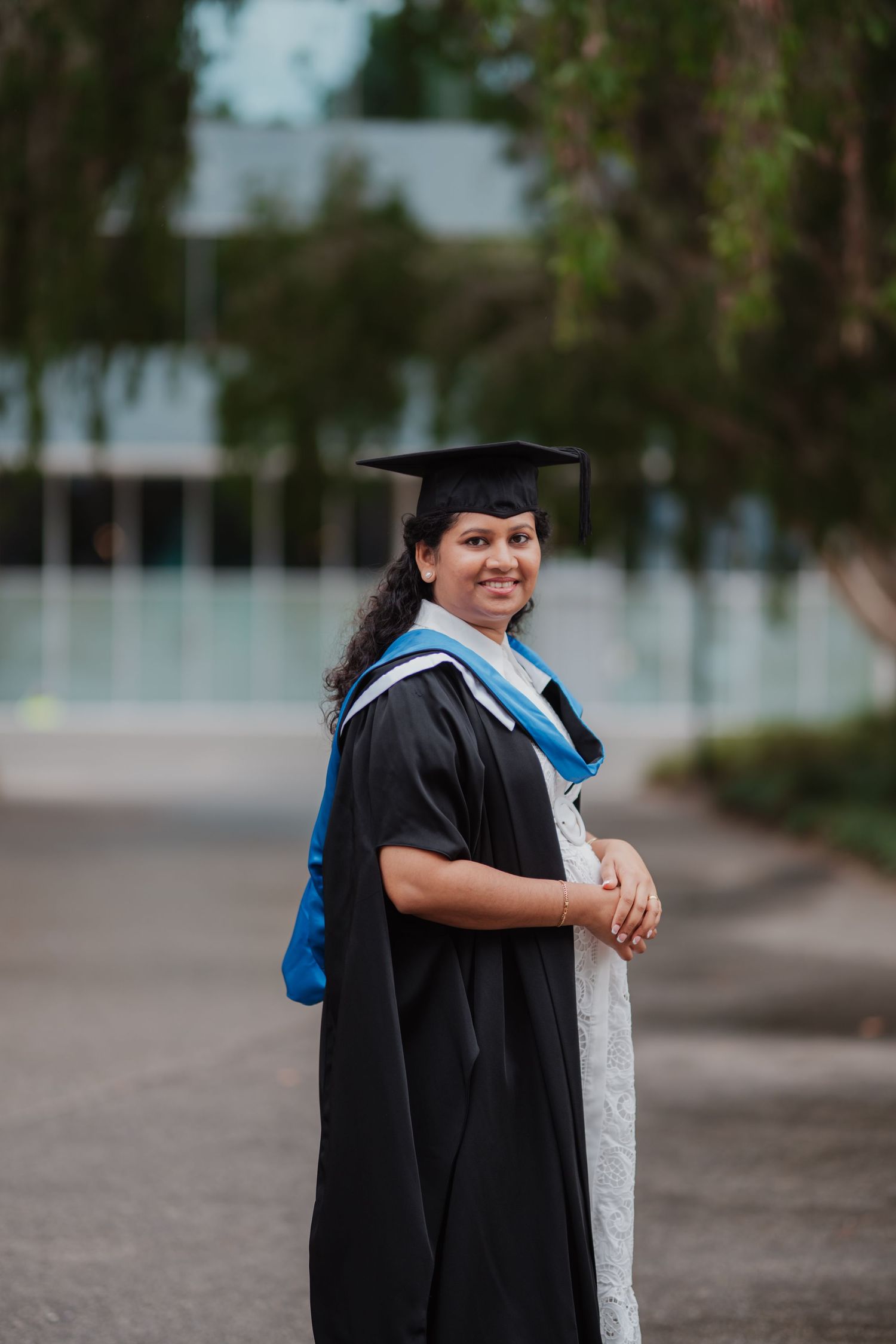 Smiling graduate in black cap and gown with blue hood poses outdoors on campus.
