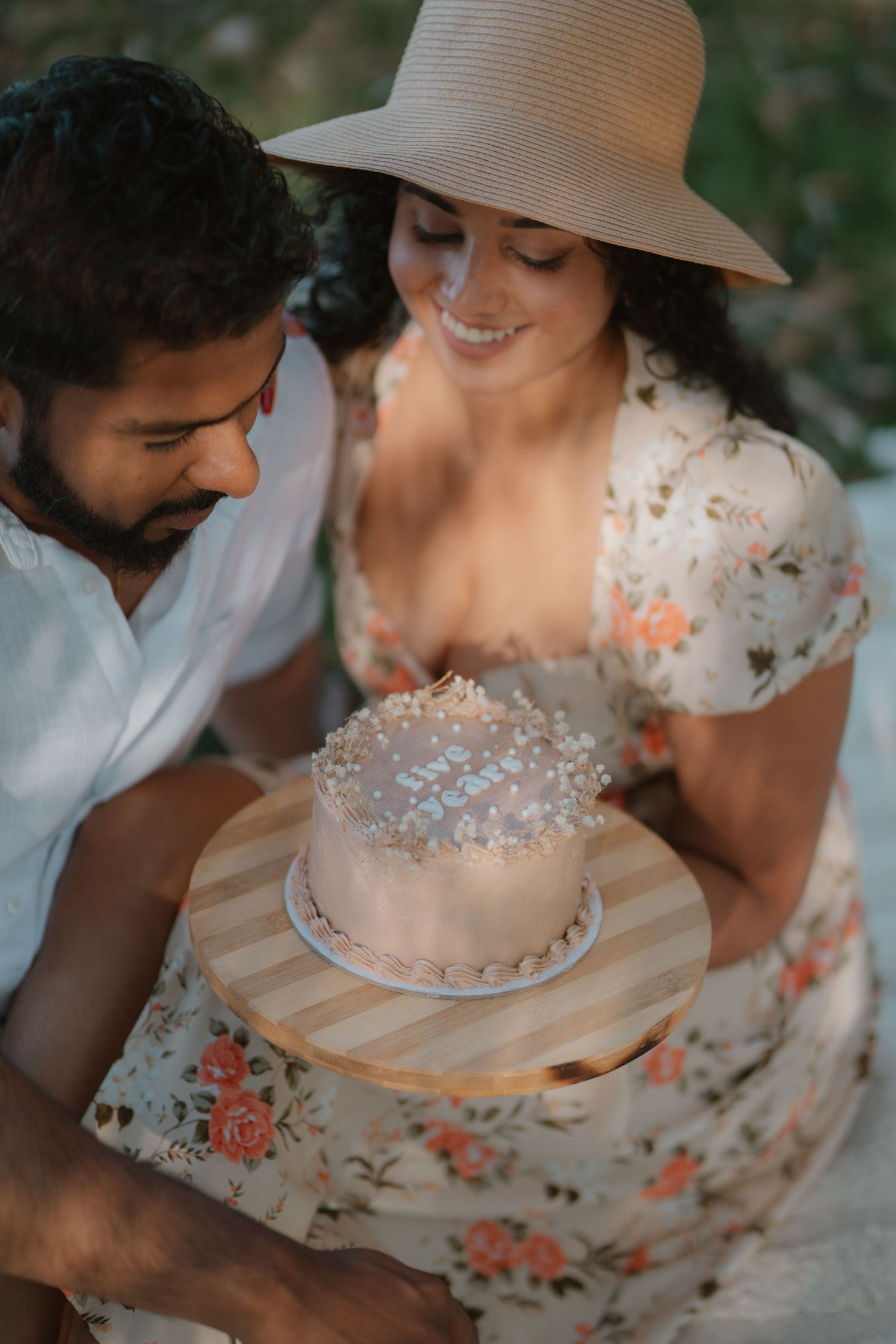 Smiling couple at outdoor picnic admires decorated birthday cake with candles in warm sunset light.