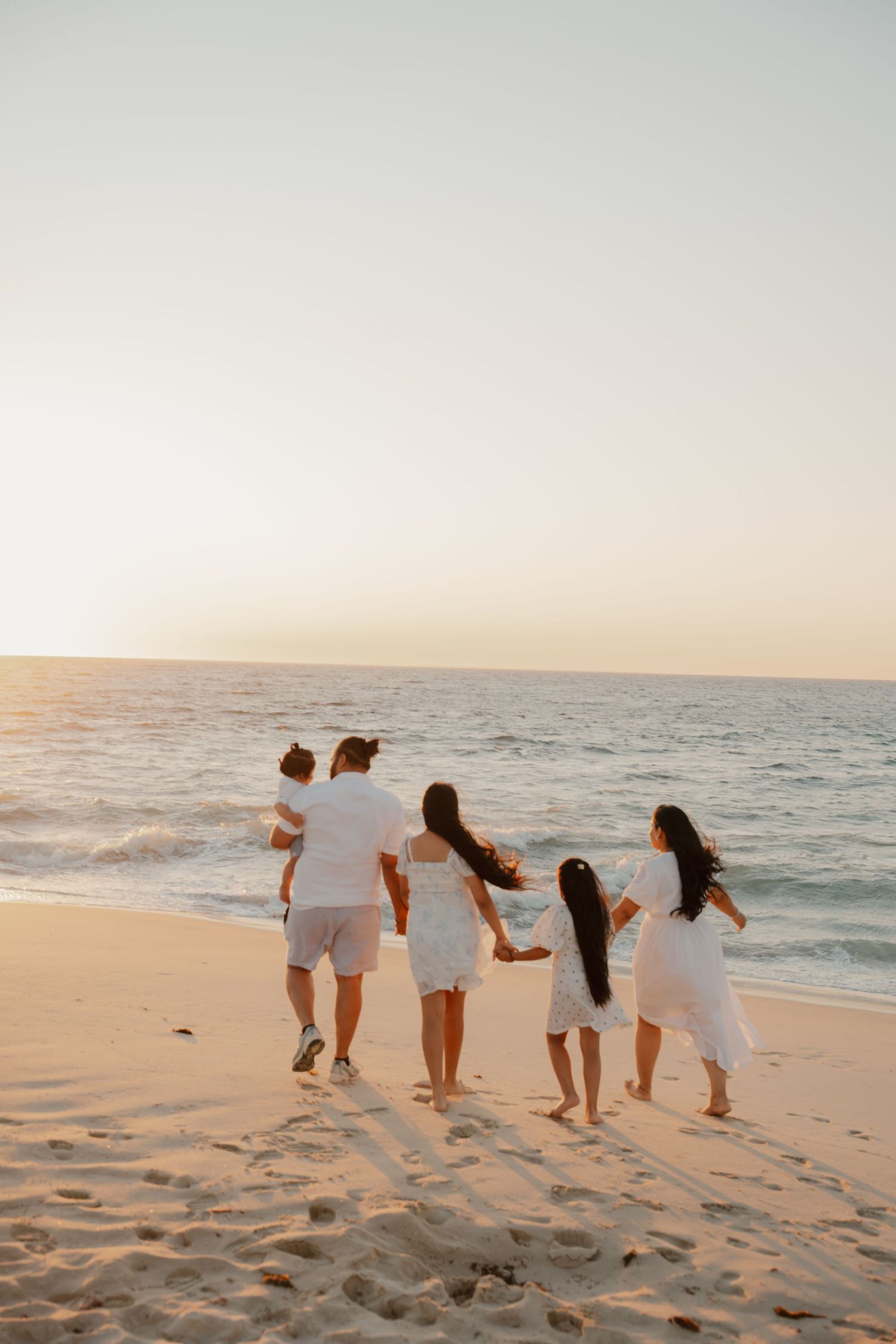 Family in white outfits walking hand-in-hand toward ocean waves at golden hour beach sunset.