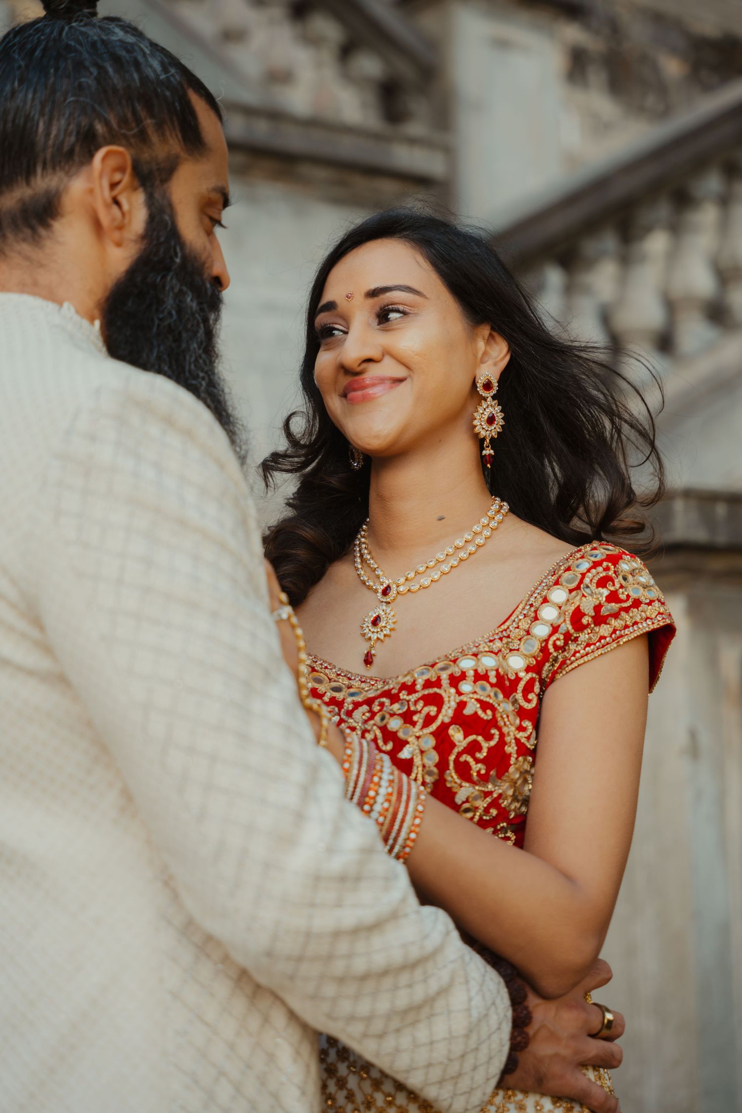 Indian bride in red and gold lehenga smiles at bearded groom in white sherwani during wedding portrait.