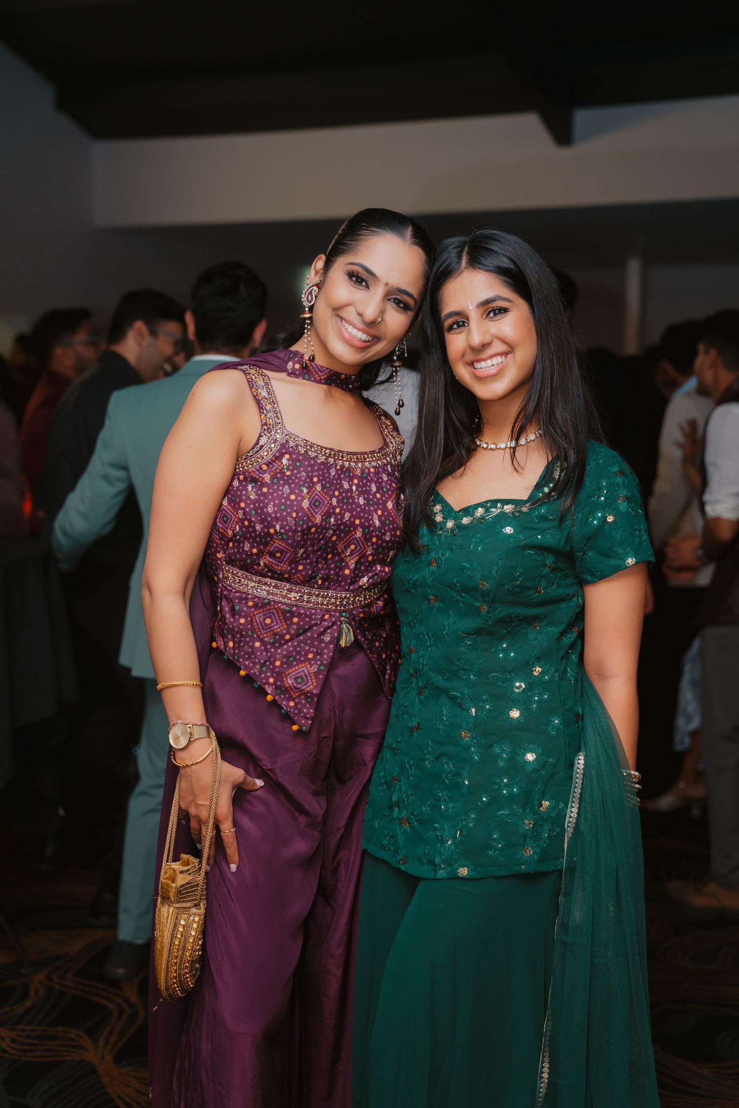Two women in purple and green Indian ethnic wear smile together at festive wedding reception.
