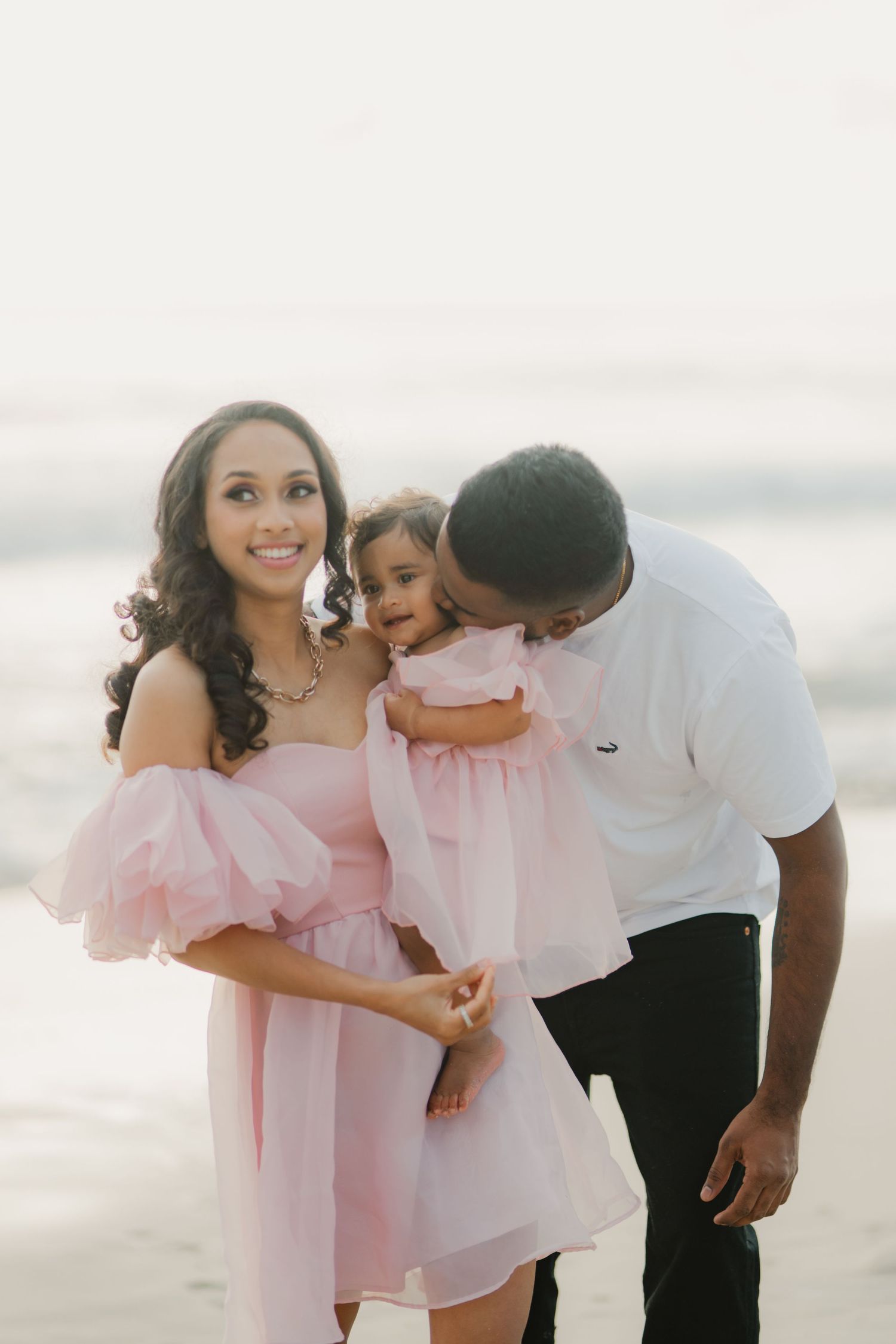 Mother smiling as father kisses toddler daughter in pink dress on beach.