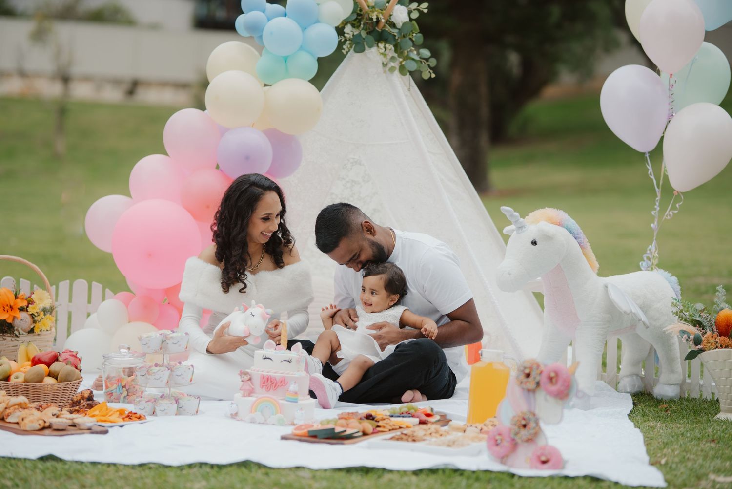 Family celebrating baby's first birthday outdoors with pastel balloons, unicorn decor, and picnic spread on white blanket.