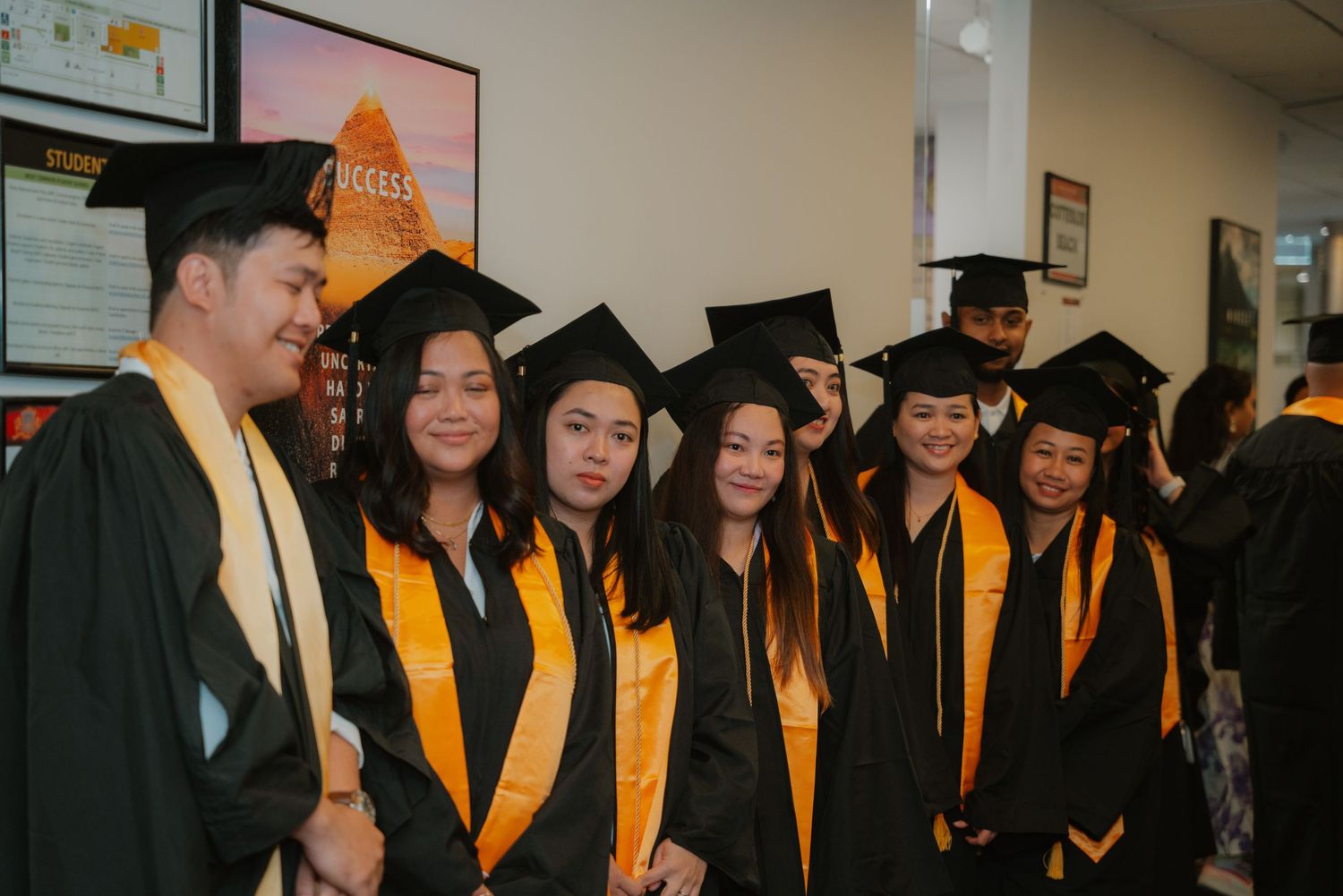 Six graduates in black and gold caps and gowns posing together for a photo indoors.
