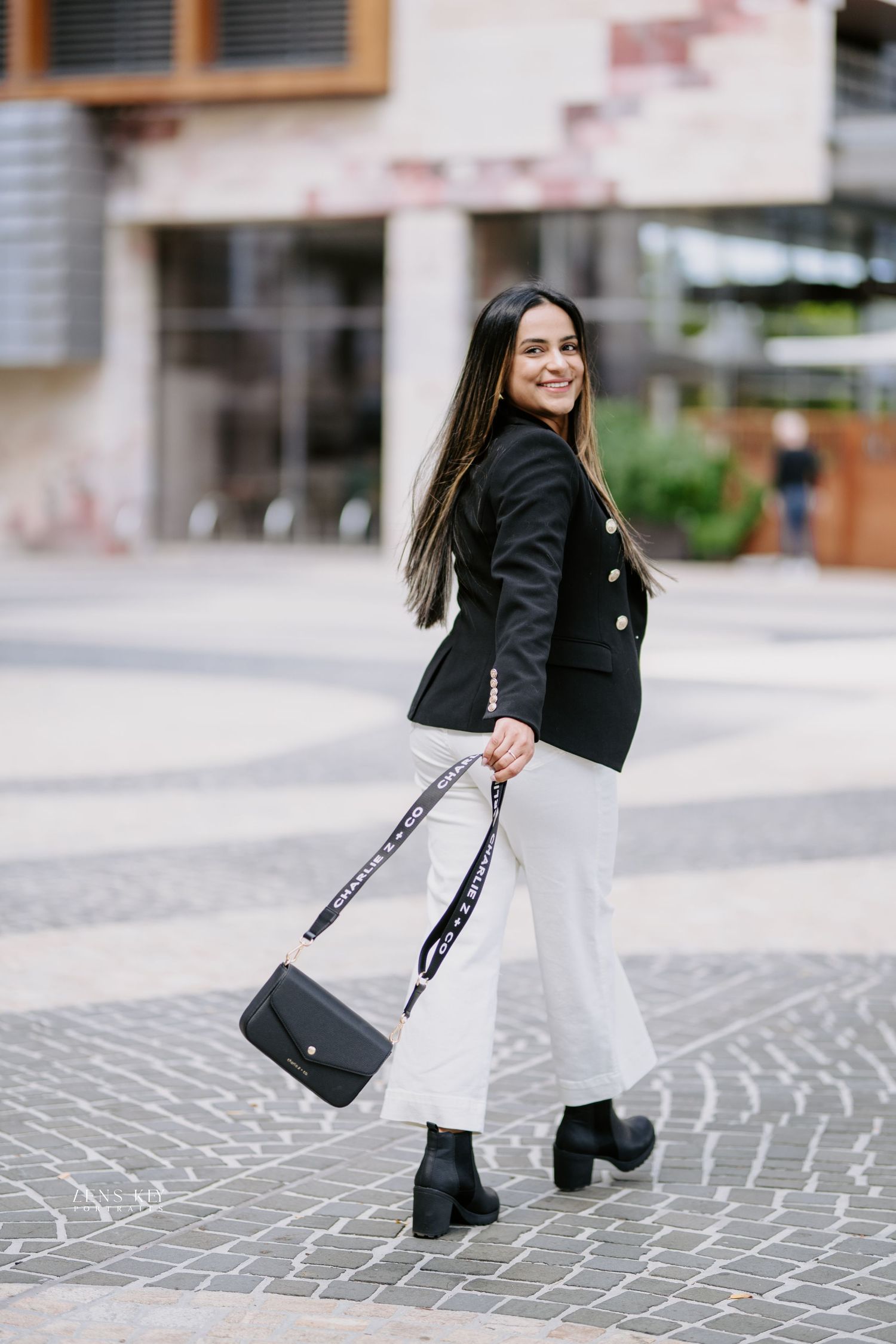 Stylish woman in black blazer and white pants carrying designer bag on city plaza.