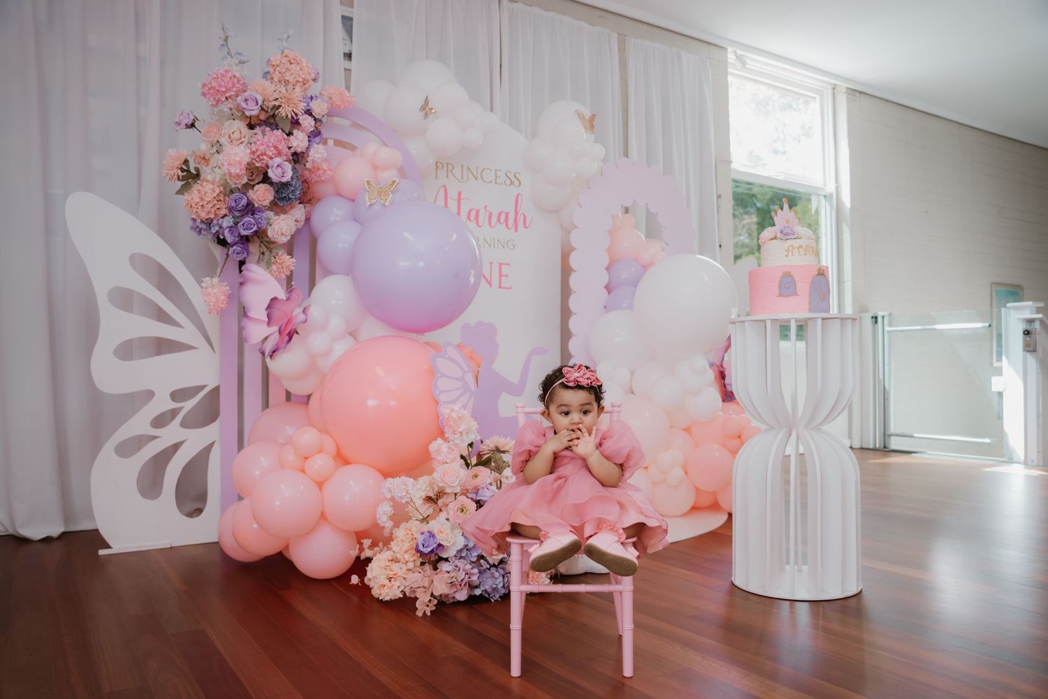 Baby girl in pink dress at butterfly-themed first birthday party with balloon arch decor.