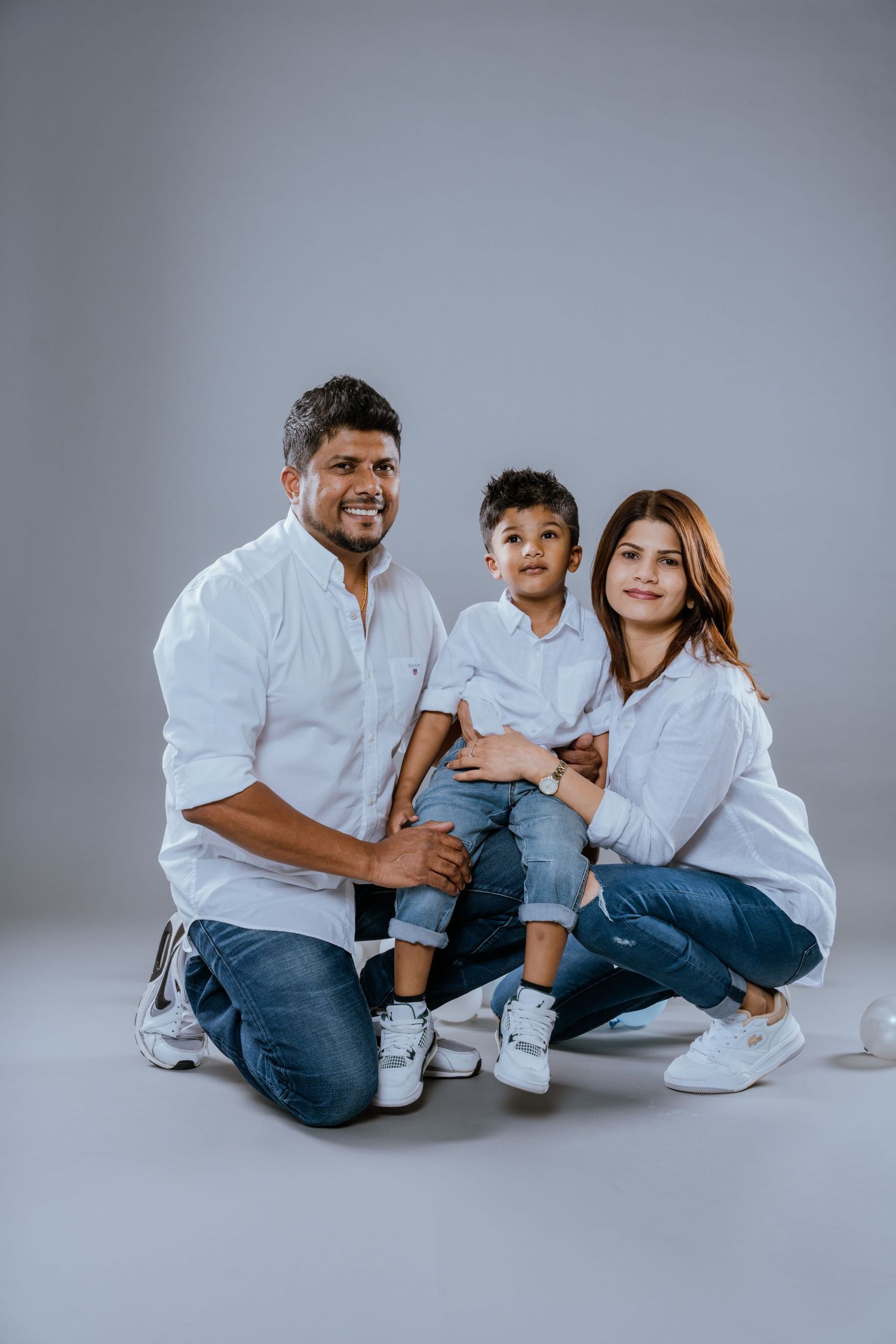 Family of three in matching white shirts and jeans pose together on a gray background.