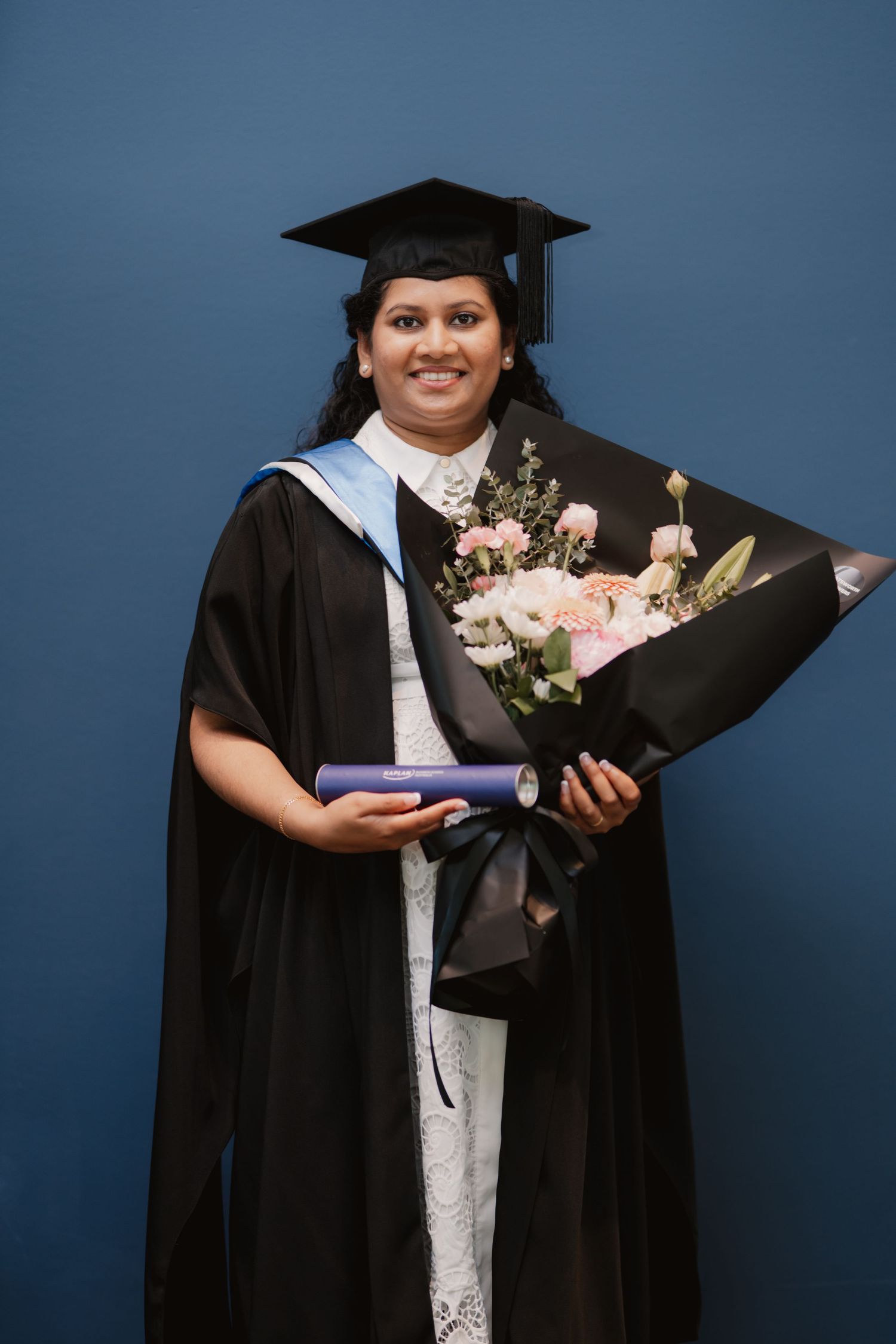 Female graduate in black cap and gown holding a diploma and bouquet of flowers against a blue wall.