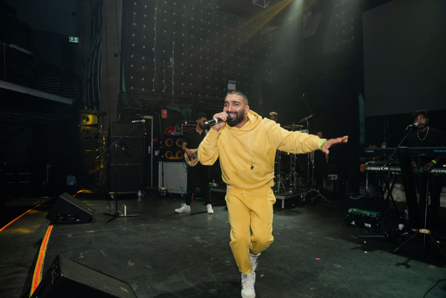 performer in yellow tracksuit singing on stage with band members in background.