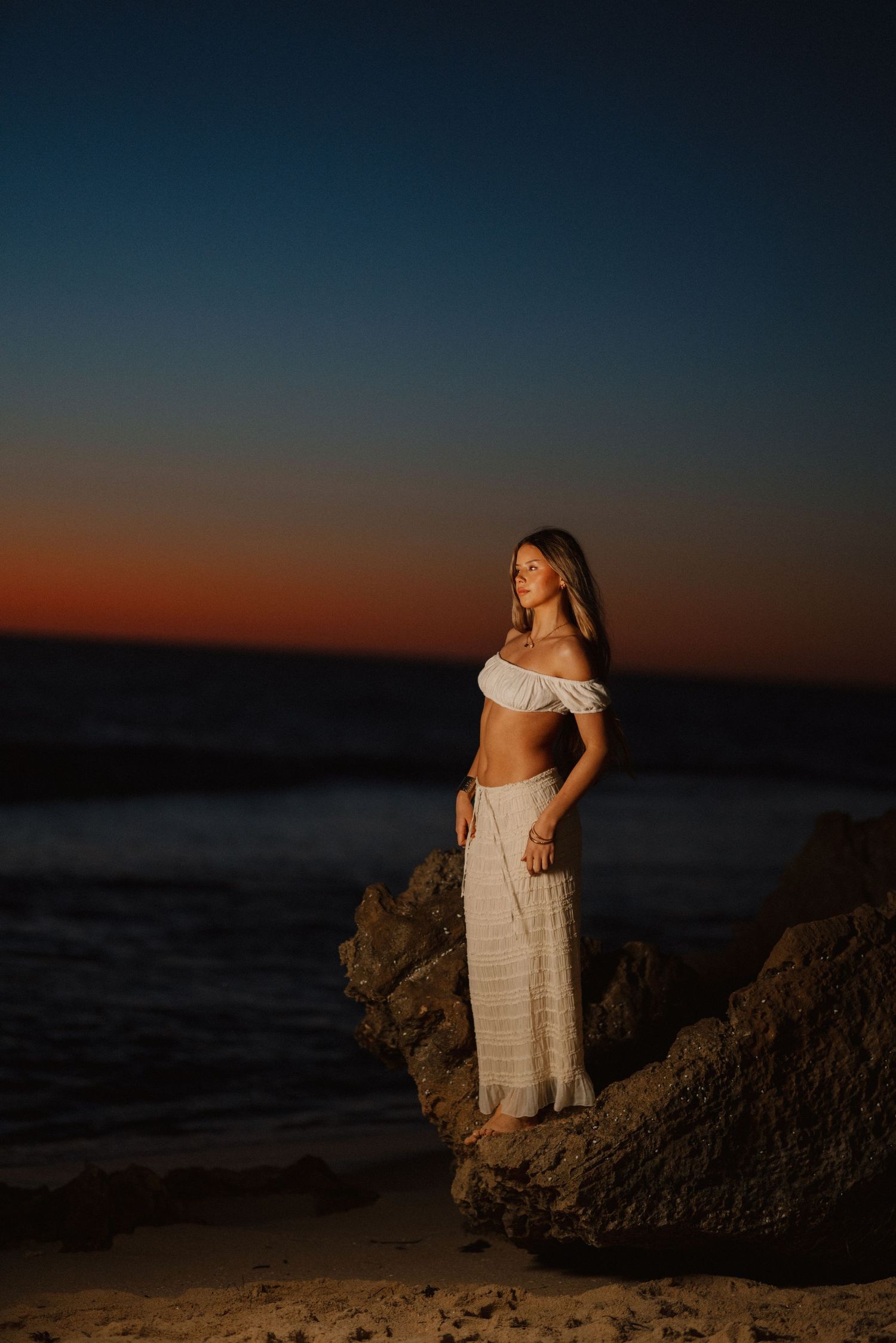 Woman in white off-shoulder crop top and skirt stands on beach rocks at dusk.