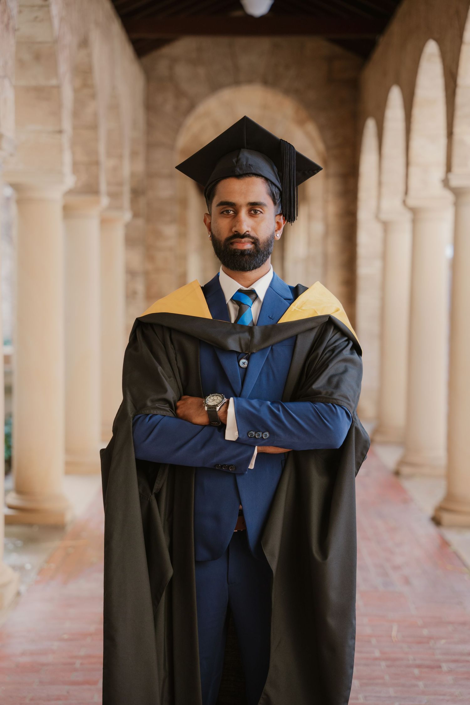 Bearded male graduate in blue suit and black graduation gown poses in arched hallway.