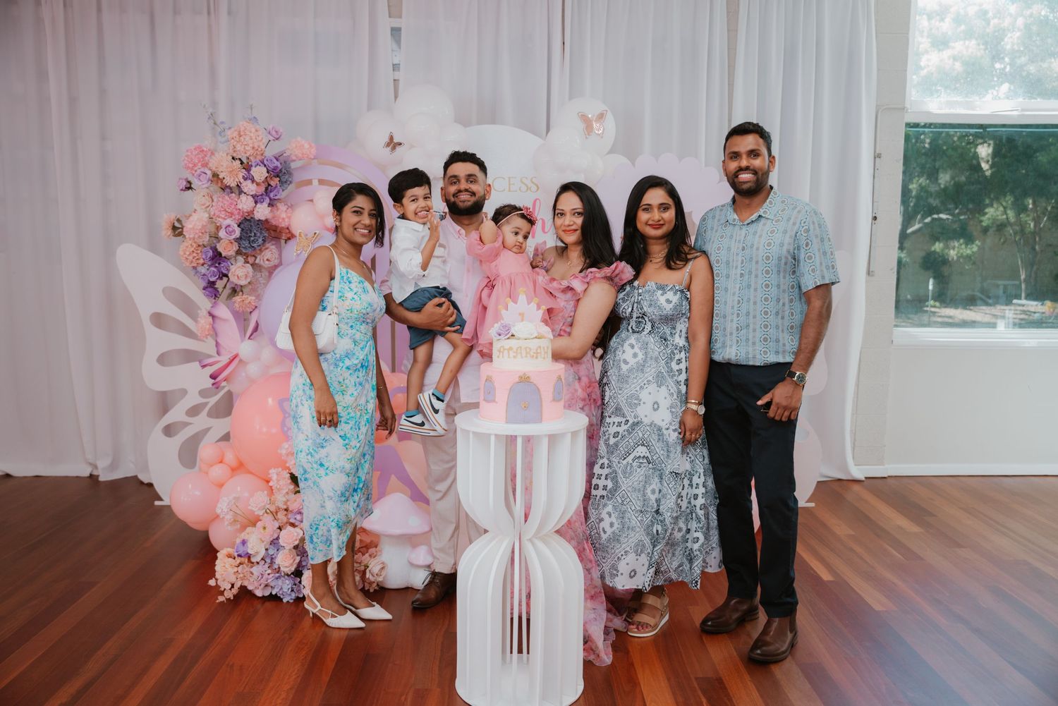 Extended family group poses with pink butterfly first birthday cake at elegant party venue.