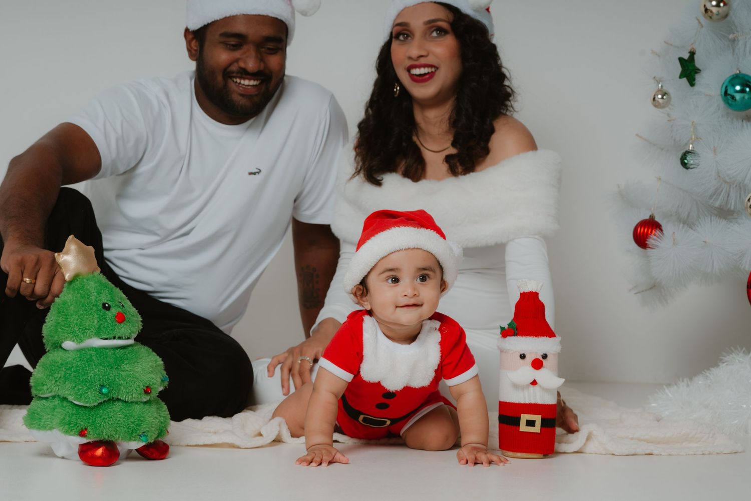 Joyful parents laughing with crawling baby in Santa outfit during a festive white Christmas photo session.