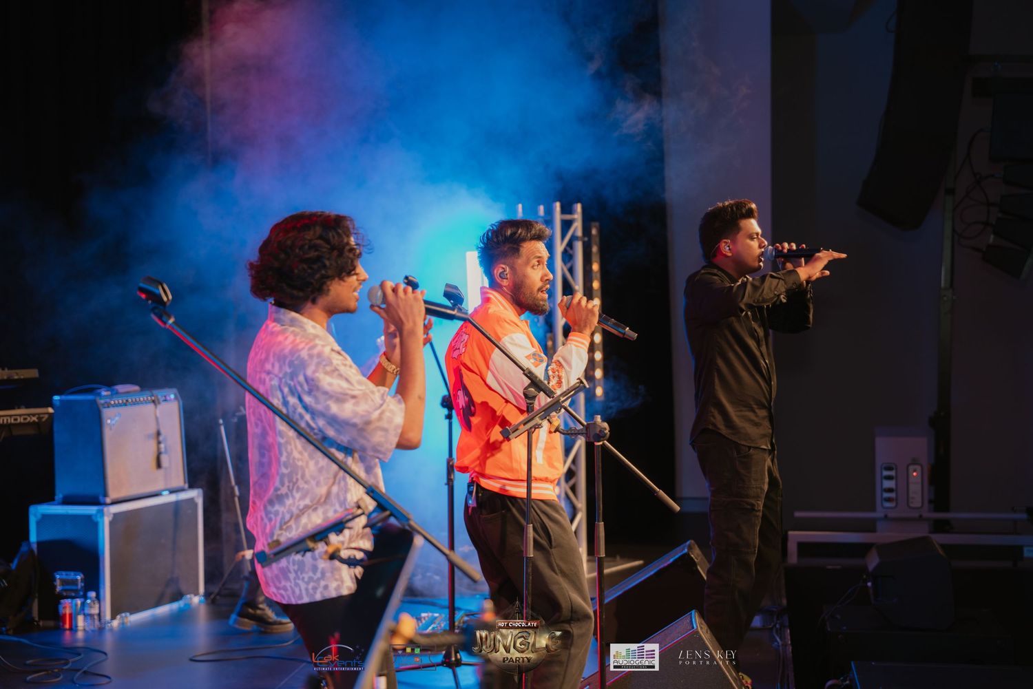 Three musicians performing on stage with blue and orange atmospheric fog lighting.