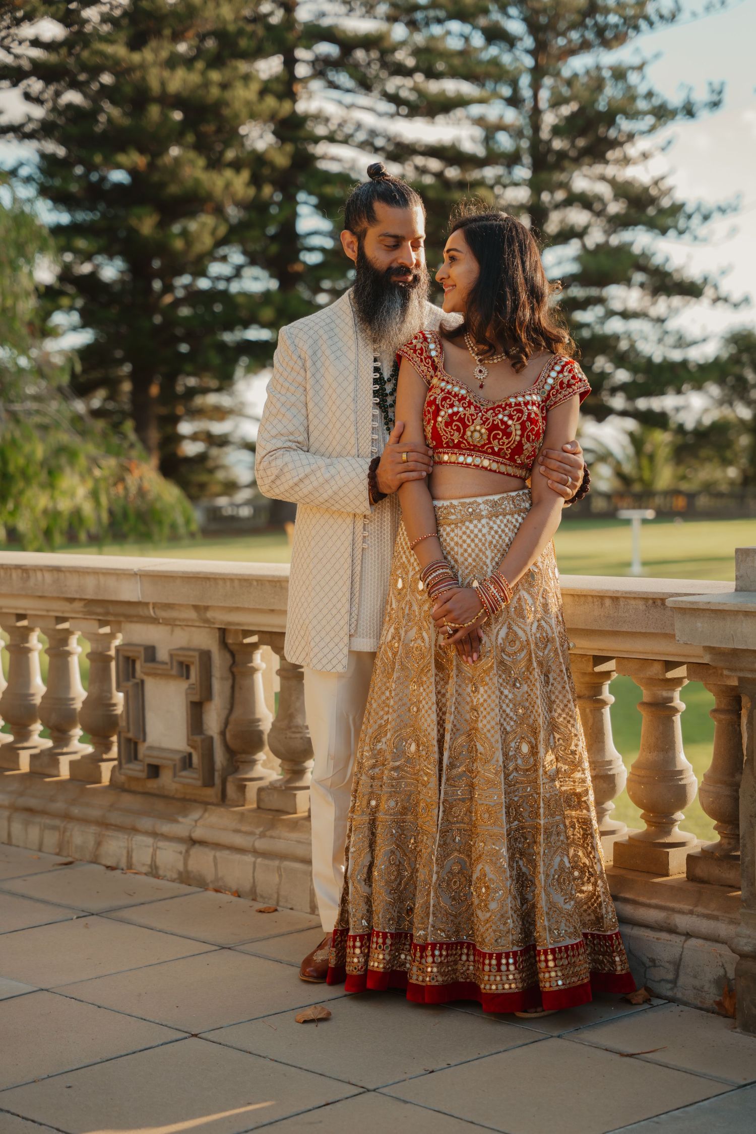 Indian bride in red and gold lehenga stands with bearded groom on stone terrace at golden hour.