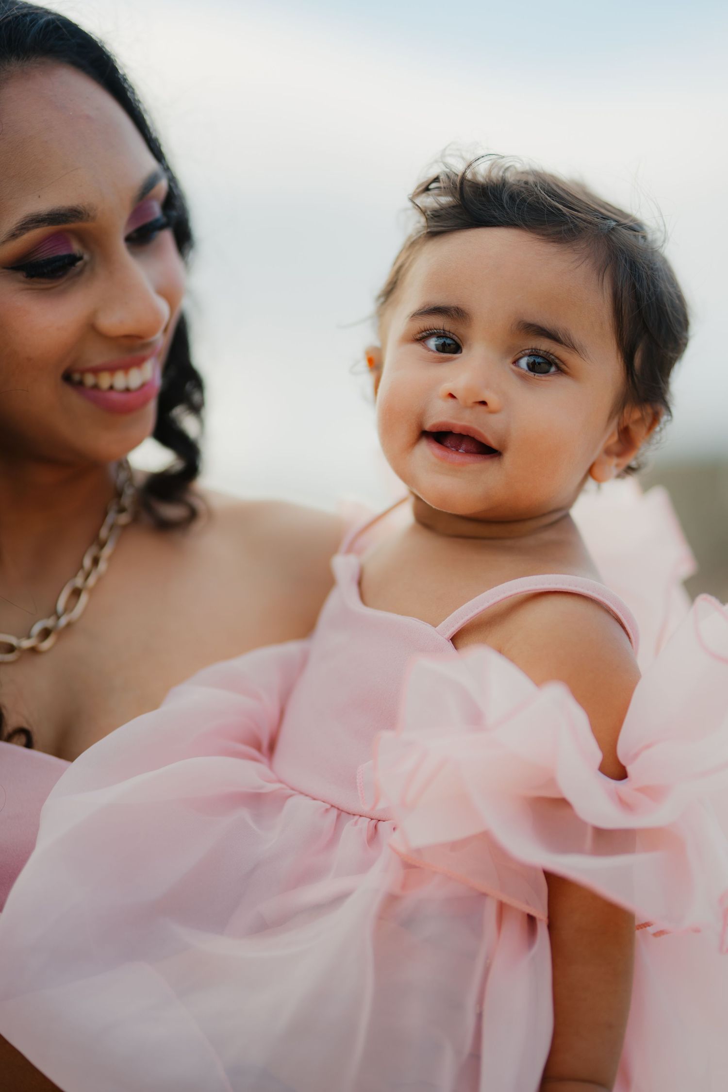 Close-up of smiling mother holding her baby girl in a pink ruffle dress outdoors.