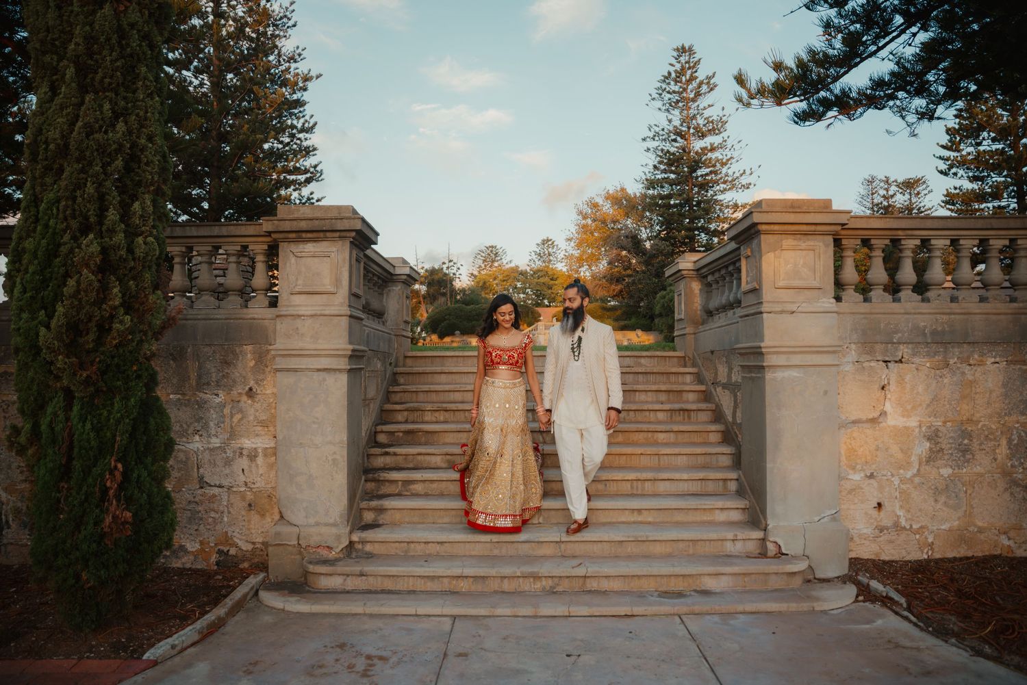 Indian bride and groom in traditional attire pose on elegant stone staircase at outdoor wedding venue.