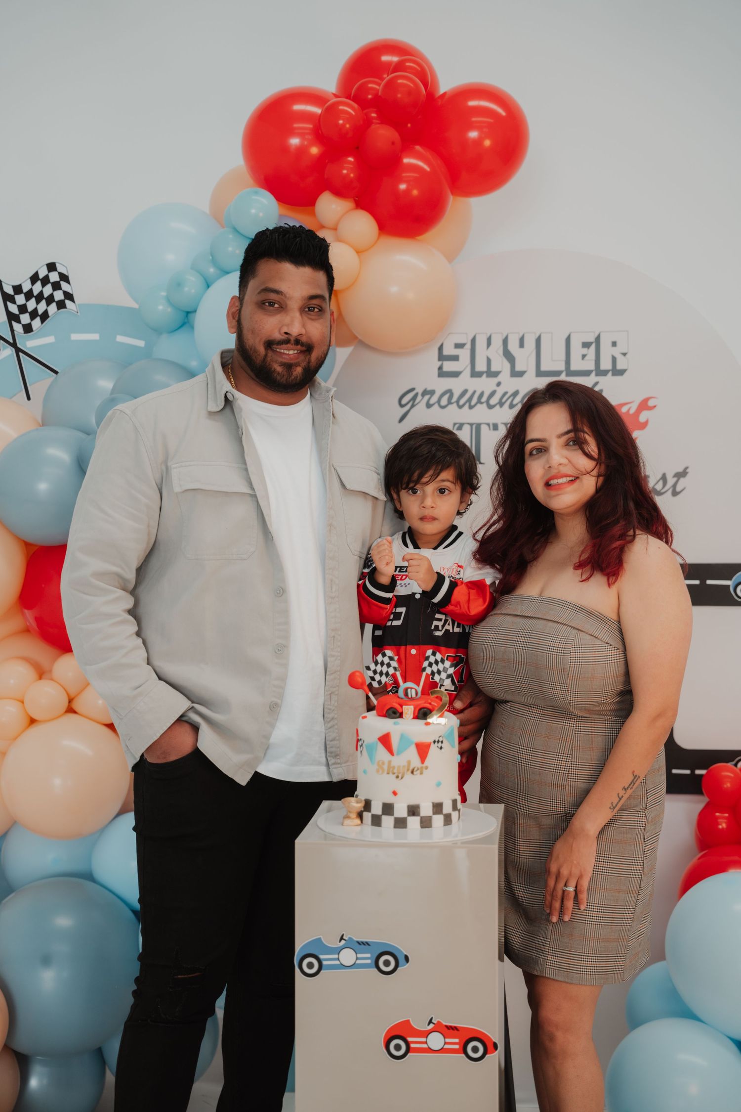 A family of three poses with a checkered race car birthday cake at Skyler's TWO fast birthday party.