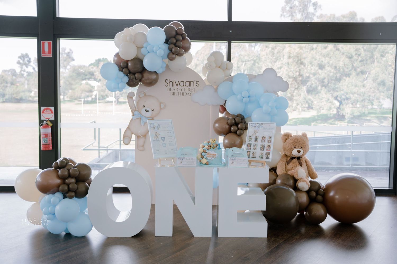 Teddy bear first birthday party decoration with blue and brown balloons and large ONE letter display.