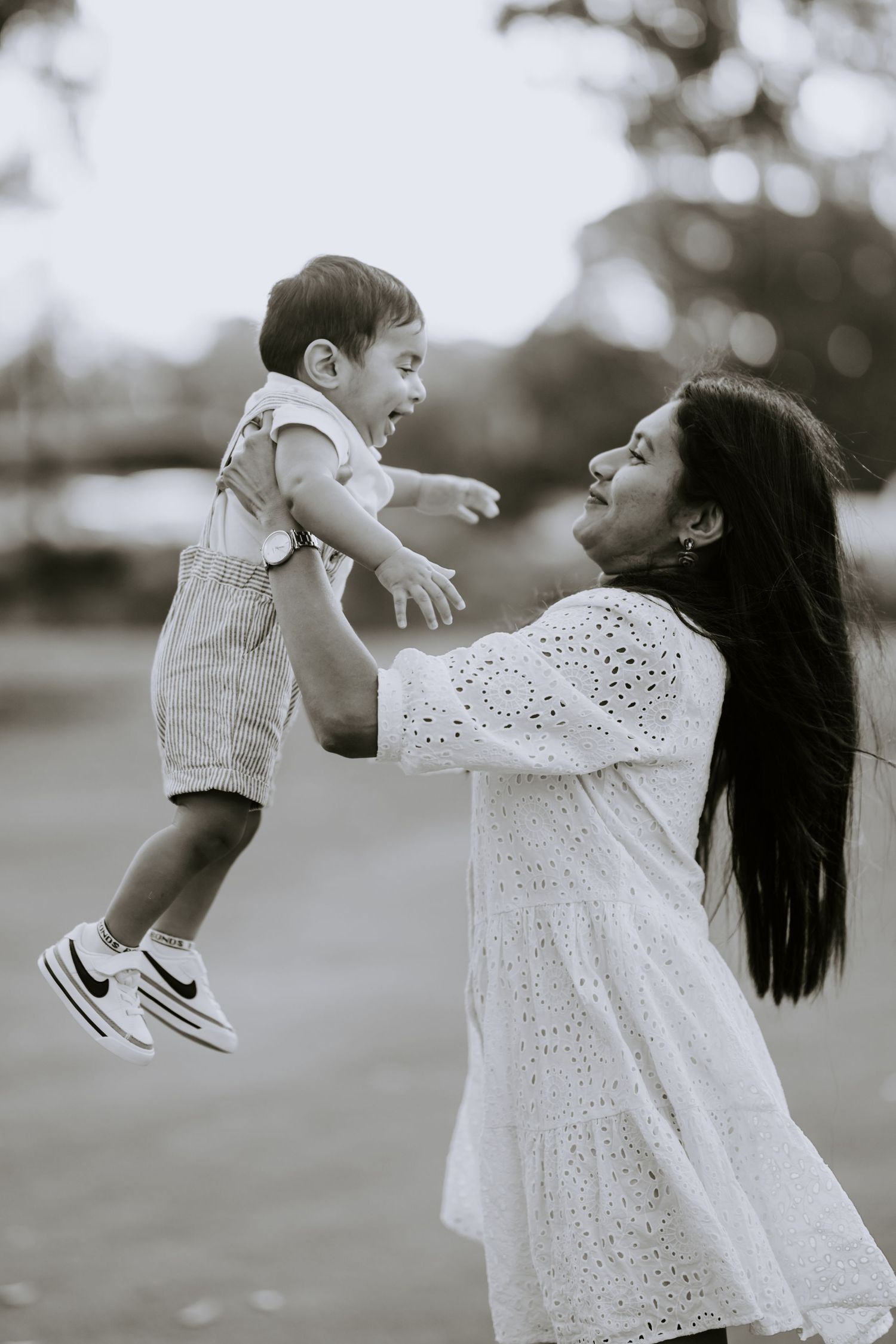 A joyful mother in white dress lifts her laughing baby outdoors in black and white photo.